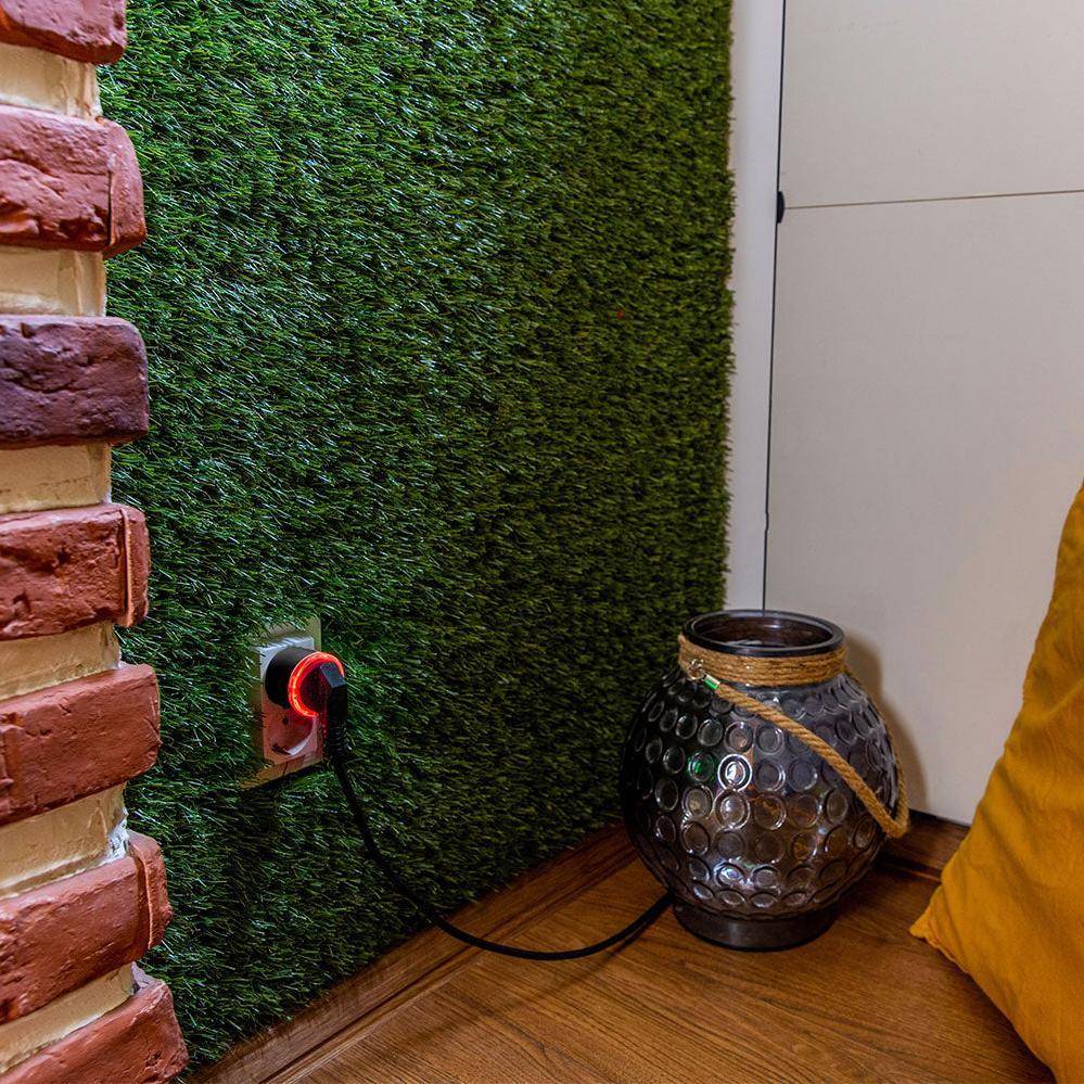 Eine Raumecke mit einem Stecker in einer Wandsteckdose, bedeckt mit grünem künstlichem Rasen; eine dekorative Vase steht auf Holzfußboden daneben.