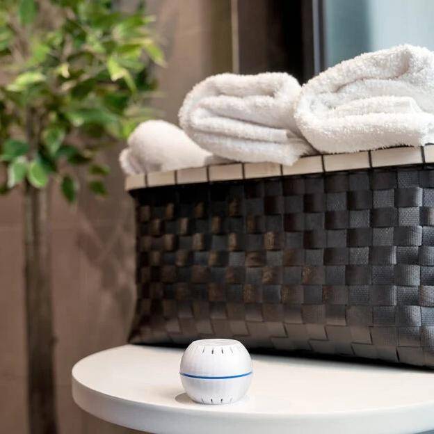 Ein geflochtener Korb mit weißen Handtüchern auf einem Tisch, daneben ein kleiner, runder Lufterfrischer, mit einer grünen Pflanze im Hintergrund.