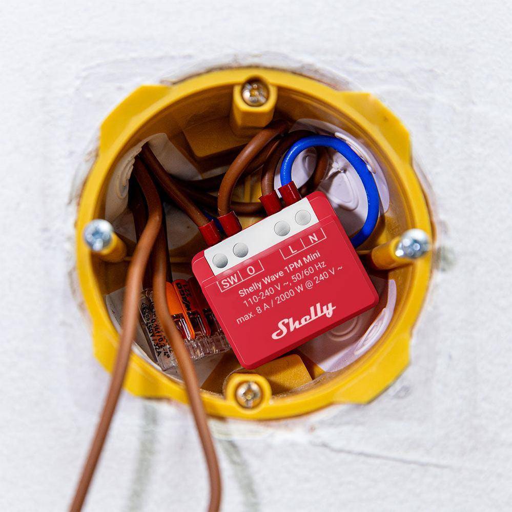 Rote Shelly-Modul mit beschrifteten Kabeln in einer kreisförmigen Wandsteckdose. Text lautet: Shelly 1PM Mini. Max: 240V~, 50/60Hz, 10A.