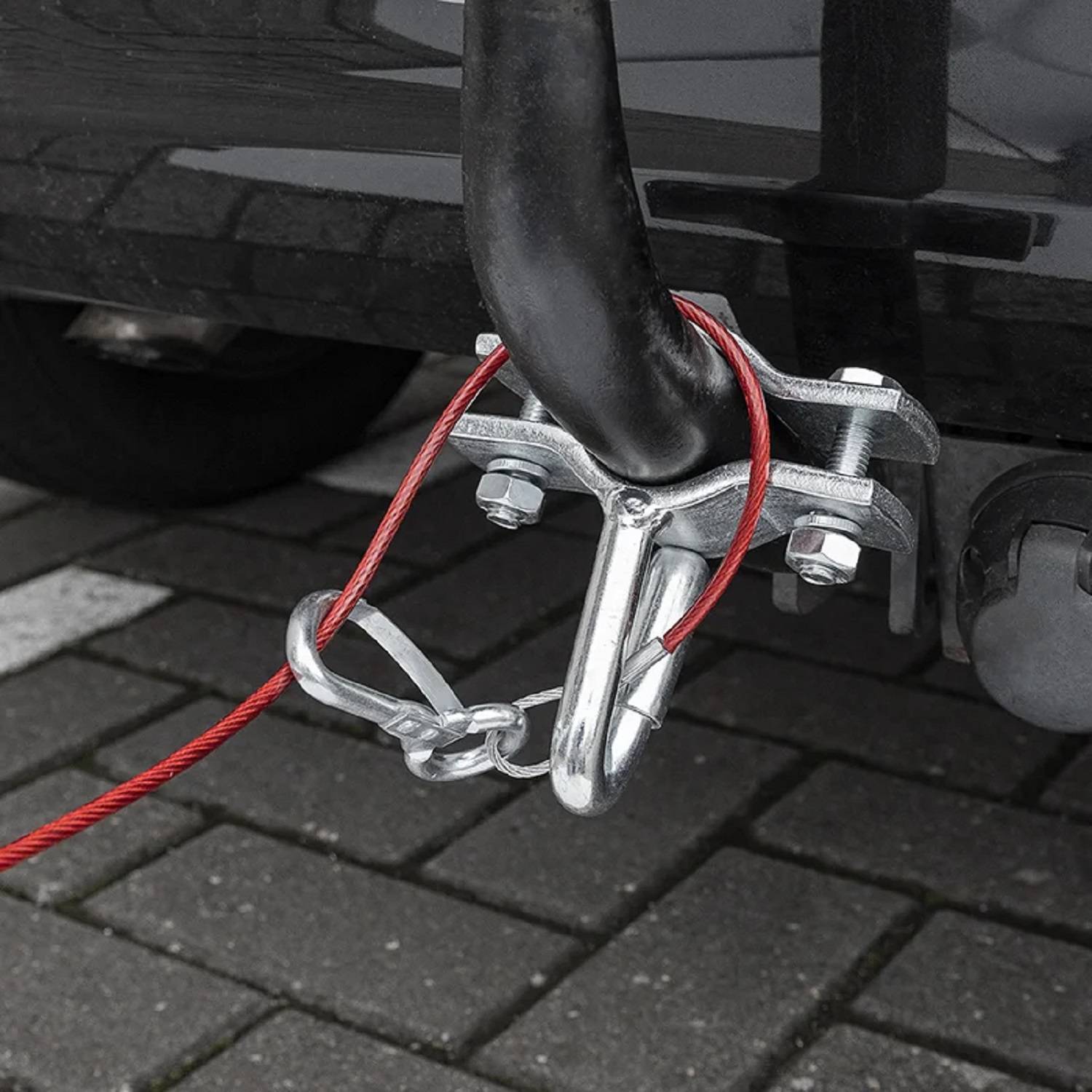 Abschlepphaken mit rotem Abschleppseil, das an einer Anhängerkupplung eines geparkten Fahrzeugs befestigt ist, auf einem grauen Pflasterstein-Parkplatz.