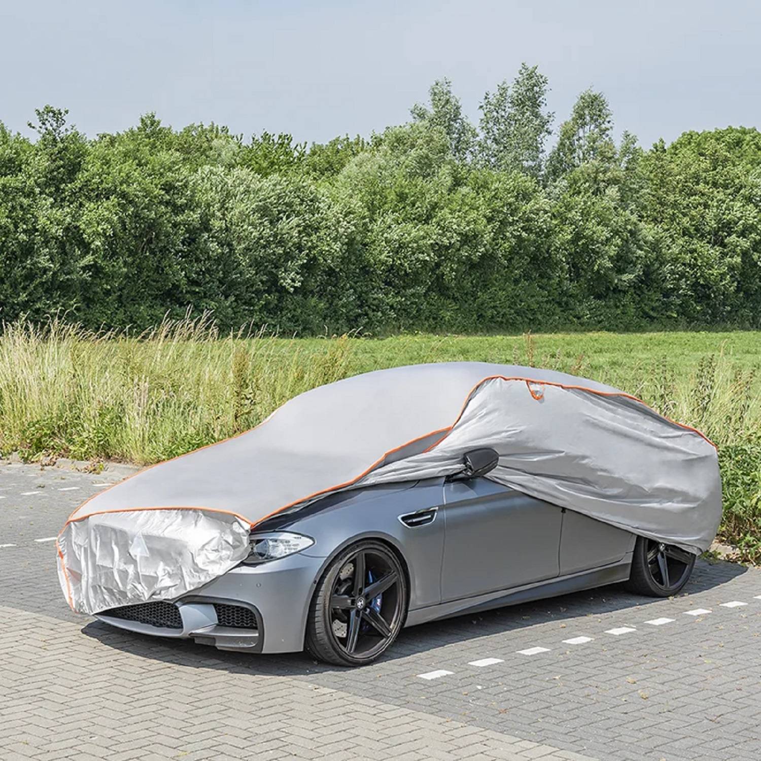Ein silberner Wagen ist auf einer asphaltierten Fläche geparkt und ist teilweise mit einer grauen Autoabdeckung verhüllt, neben einem Rasenfeld mit Bäumen im Hintergrund.