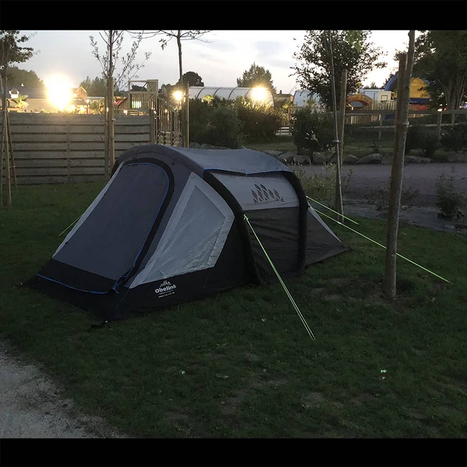 Ein kleines Zelt ist auf Gras aufgestellt, mit Seilen, die an nahegelegenen Bäumen verankert sind, umgeben von einem Campingplatz in der Abenddämmerung mit Lichtern in der Ferne.