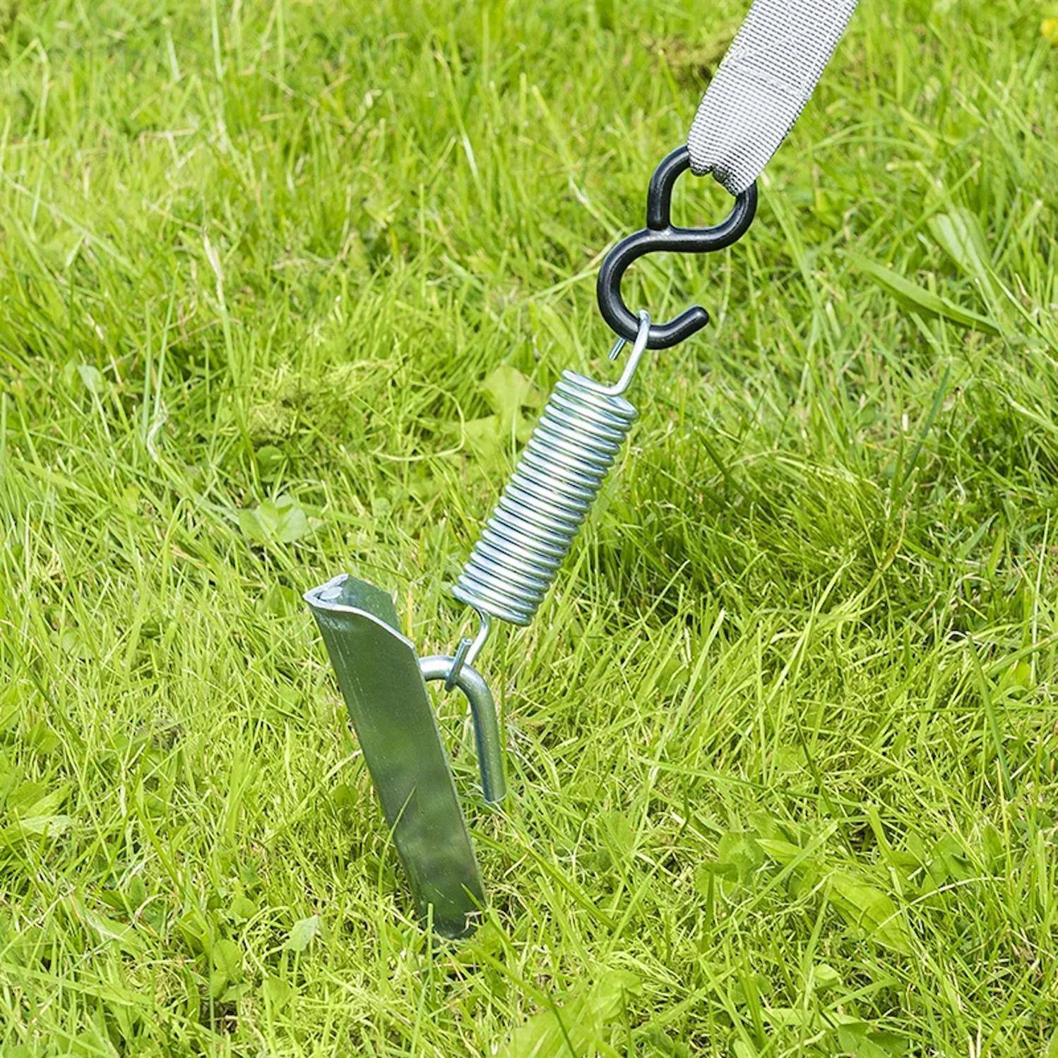 Ein Metallpfahl zur Hundebefestigung ist in einem Grasbewuchs verankert, mit einer Schraubenfeder und Karabiner, die an einer Leine befestigt sind, was eine Außen-Heimtierkontrolle andeutet.
