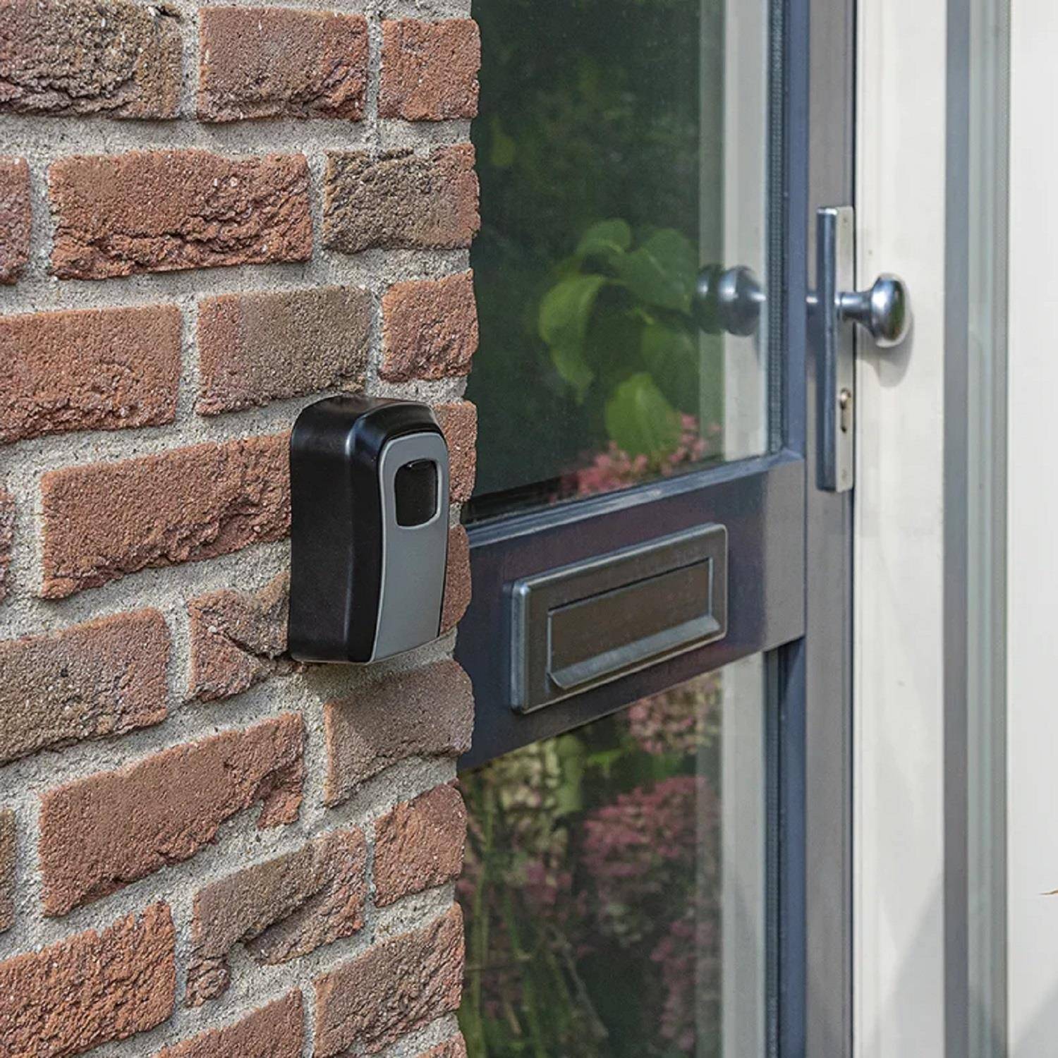 Eine schwarze Türklingel mit Kamera ist an einer Ziegelwand neben einer Glastür mit Briefschlitz montiert, umgeben von Grünpflanzen im Außenbereich.