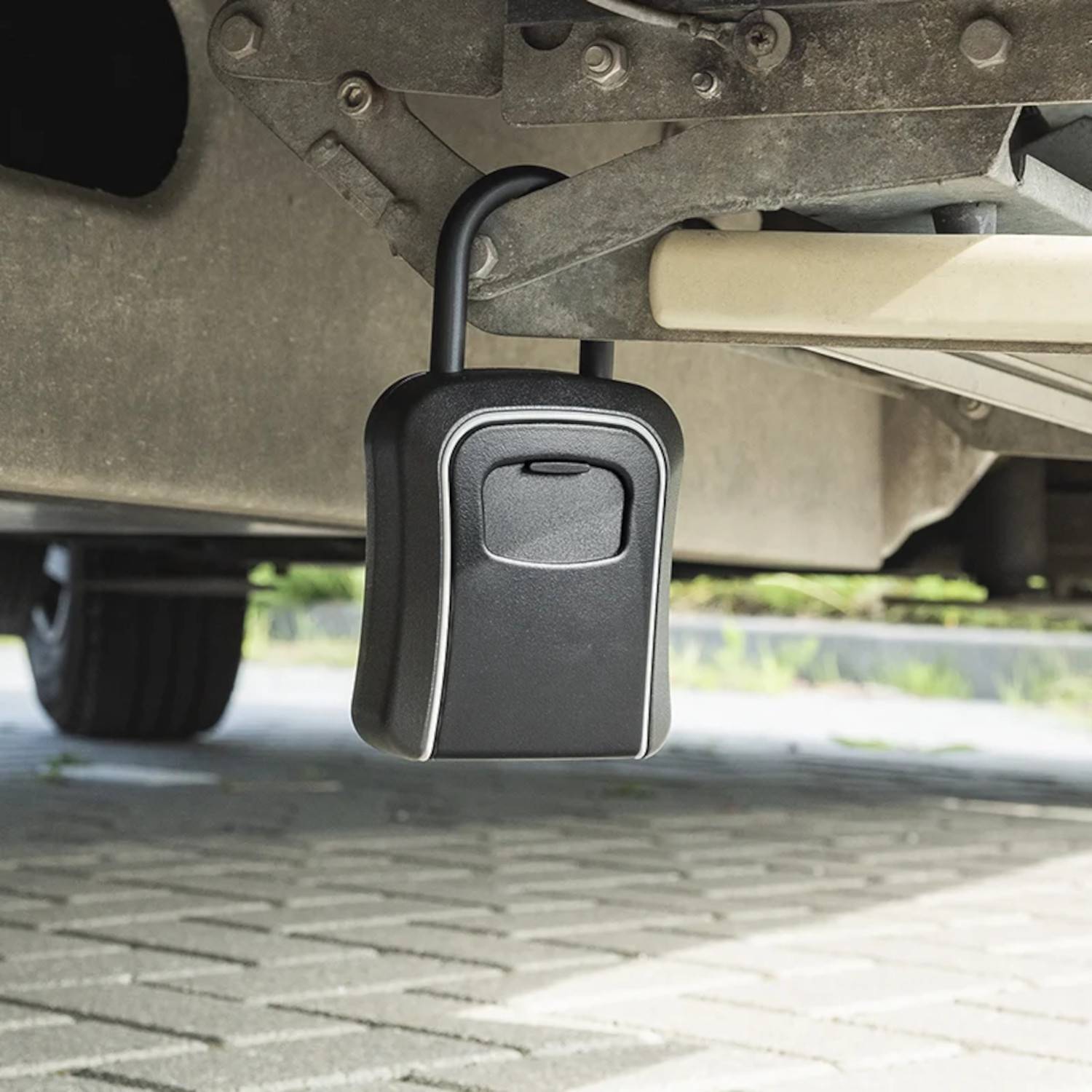 Eine schwarze Transportbox ist an der Unterseite eines Fahrzeugs befestigt und ruht auf einer asphaltierten Oberfläche, während im Hintergrund Vegetation zu sehen ist.