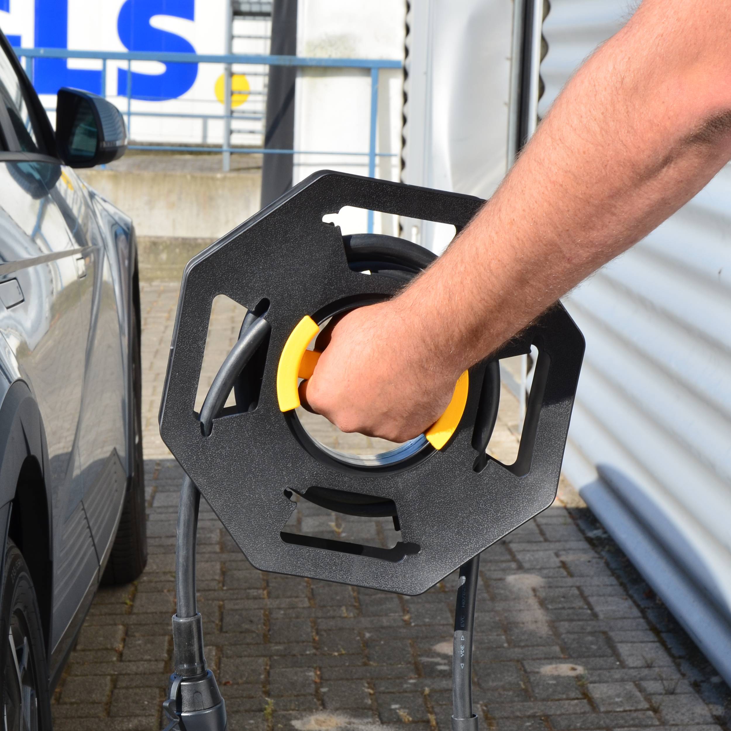 Eine Person steckt ein Elektroauto an einer Ladestation im Freien an, wobei sie ein Ladekabel in der Hand hält.