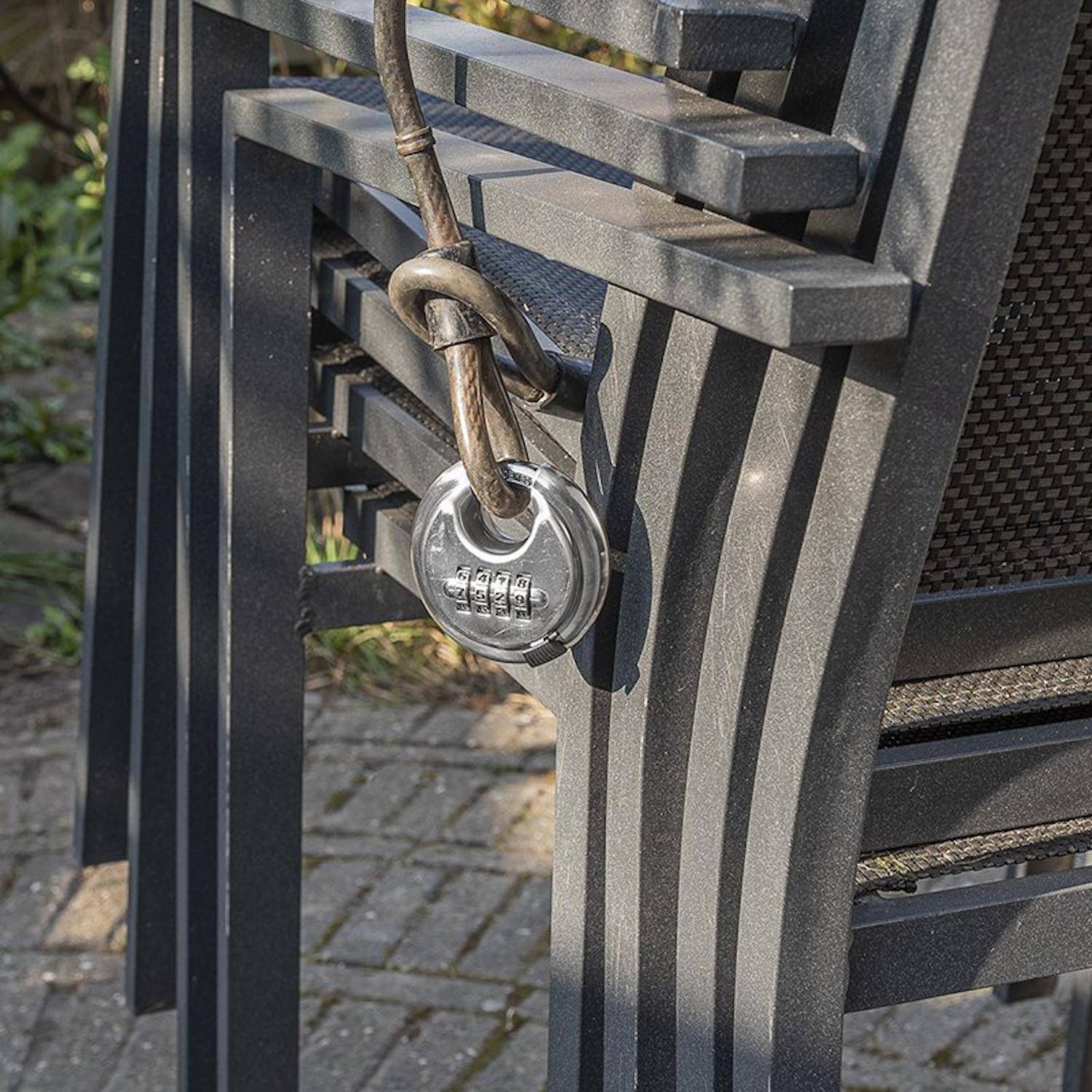Ein Metallkabel, das mit einem Zahlenschloss gesichert ist, befestigt einen Stapel von Außenstühlen auf einer Terrasse und deutet auf Sicherheit und Diebstahlprävention hin.
