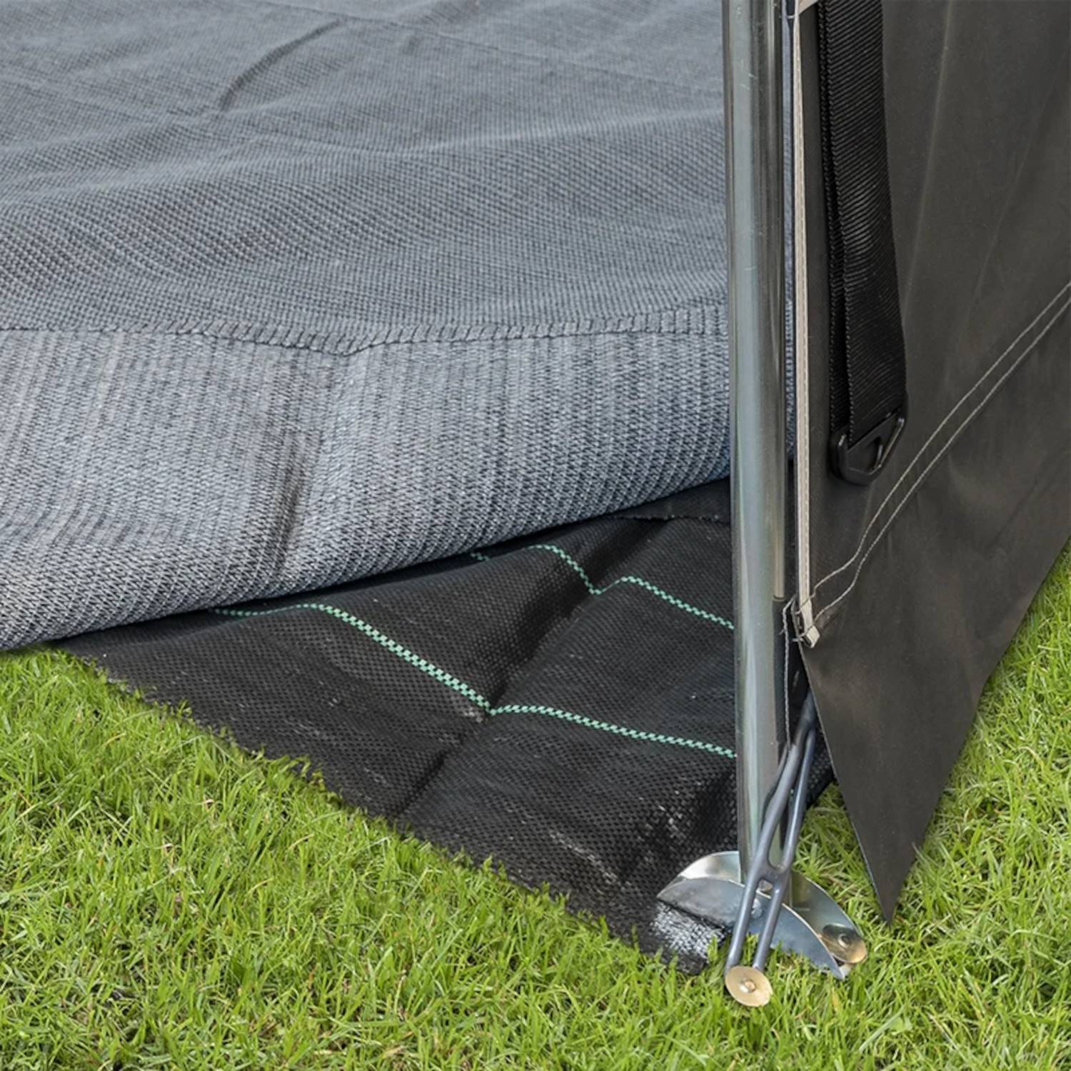 Ein grauer Outdoor-Teppich mit gewebtem Muster liegt auf Gras und bedeckt teilweise eine schwarze Matte darunter. Eine Metallstruktur steht in der Nähe.