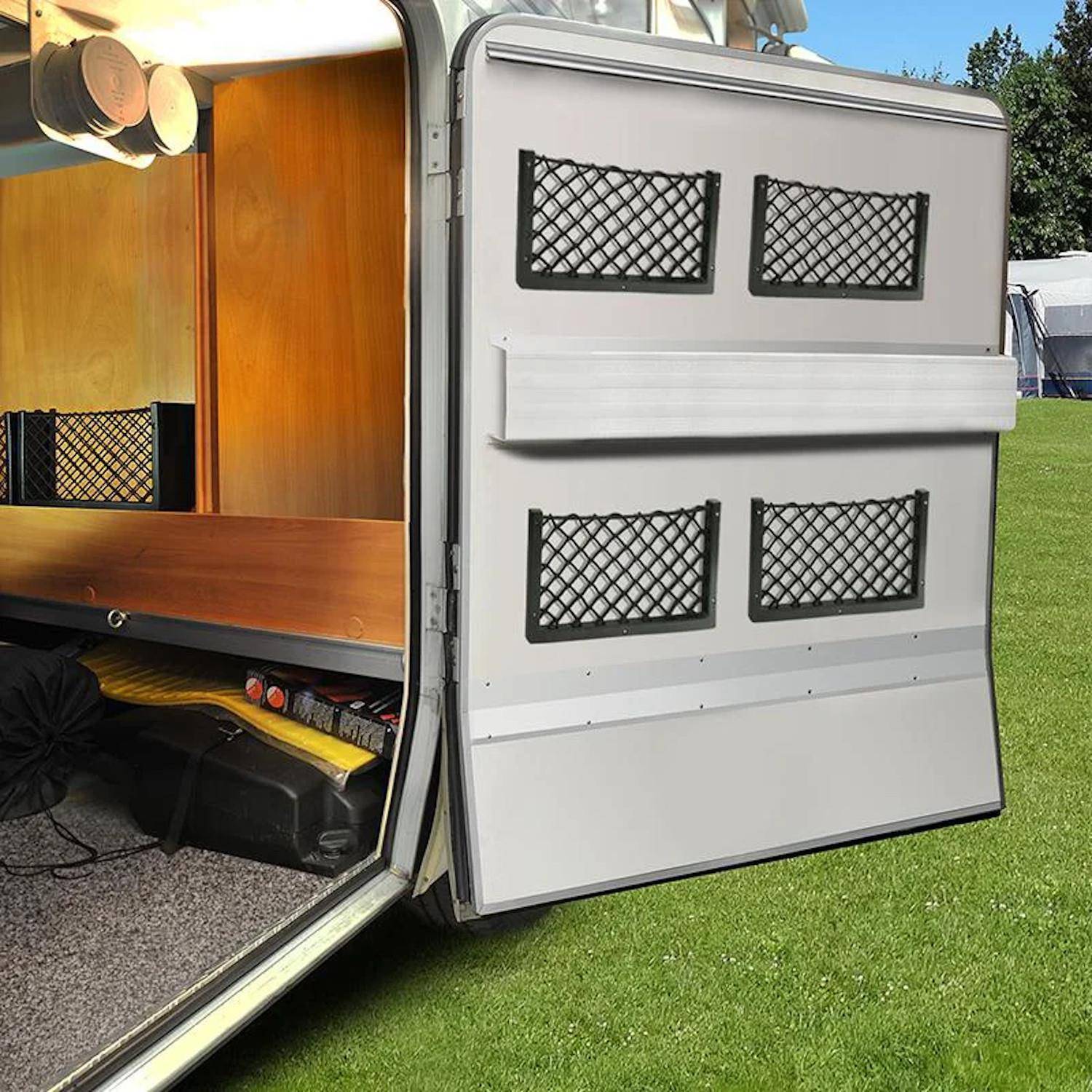 Das Bild zeigt die geöffnete Hecktür eines Fahrzeugs, die einen Innenraum mit Metallverkleidung und Netztaschen offenbart, geparkt auf Gras.