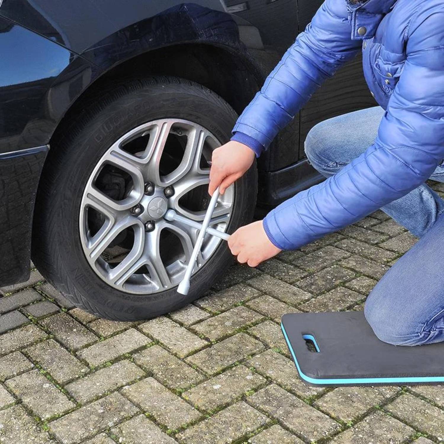 Eine Person in einer blauen Jacke verwendet einen Schraubenschlüssel, um die Radmuttern eines Autoreifens auf einer Kopfsteinpflasteroberfläche festzuziehen, was Reifenwartung signalisiert.