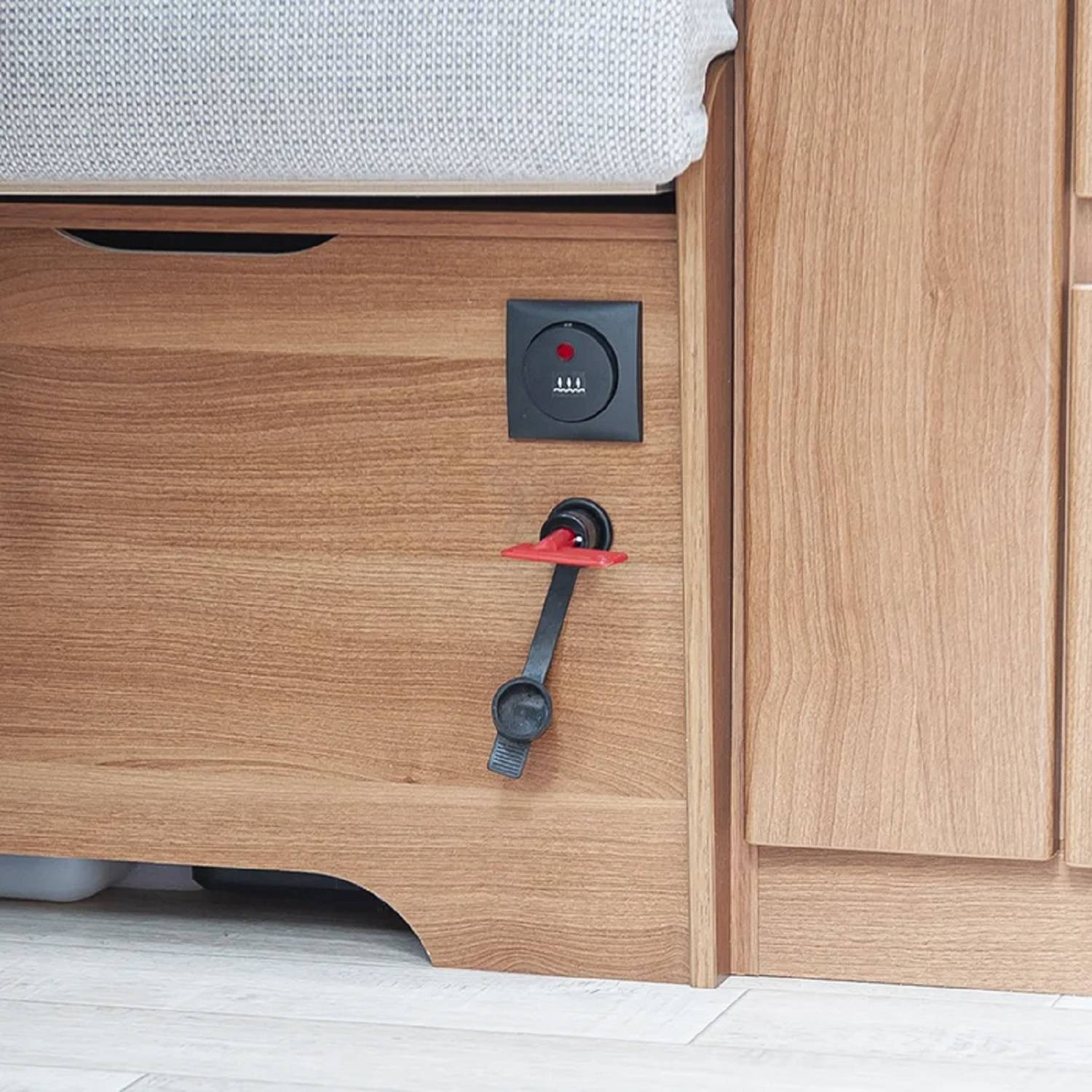 Ein Holzschrank mit einem schwarzen runden Zifferblatt und rotem Einstellknopf, neben einem grauen gepolsterten Sitz, auf einem hellen Holzboden.