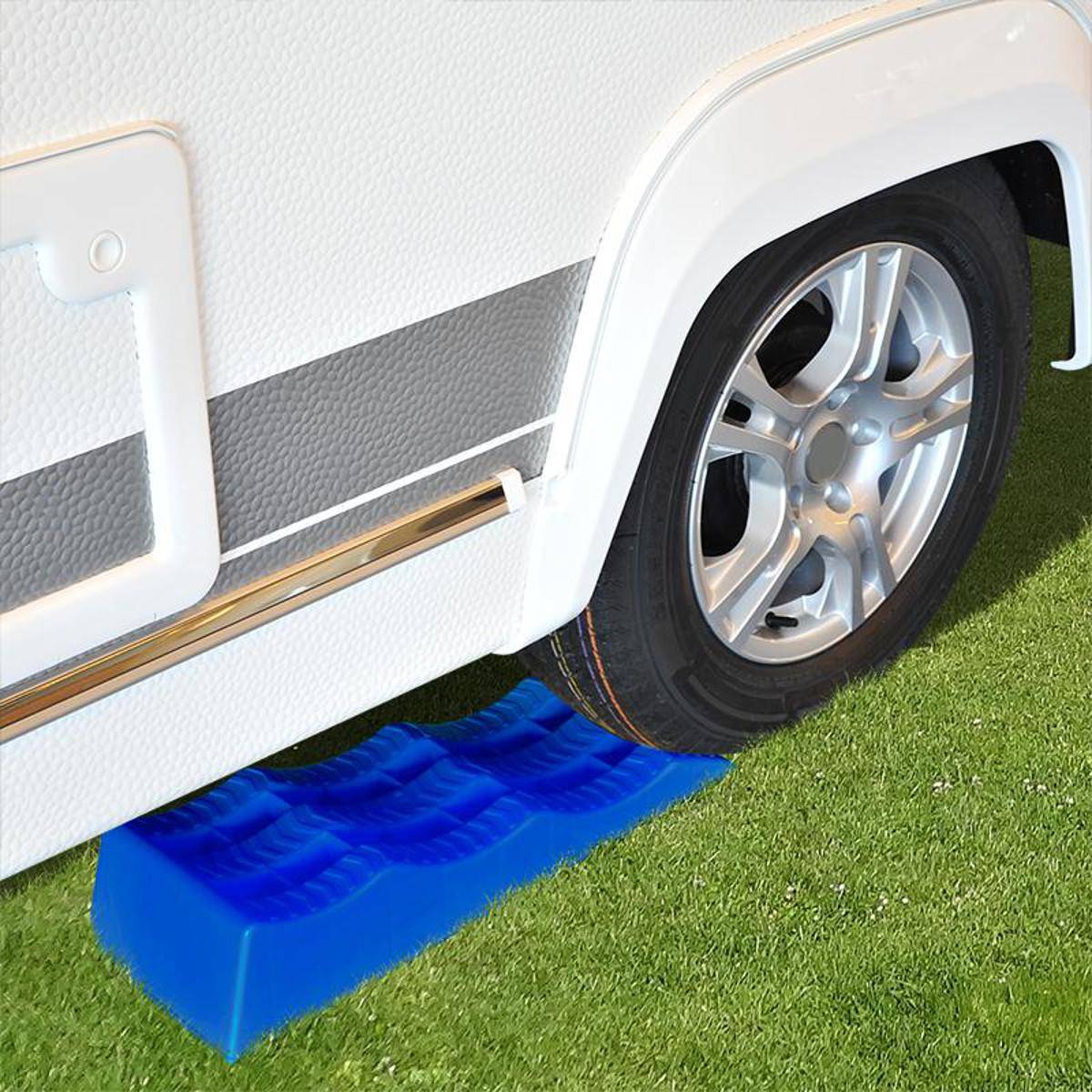 Ein Autoreifen ruht auf einem blauen Nivelierblock auf Gras, was darauf hindeutet, dass er geparkt oder stabilisiert ist, möglicherweise auf einem Campingplatz oder in einer Outdoor-Umgebung.