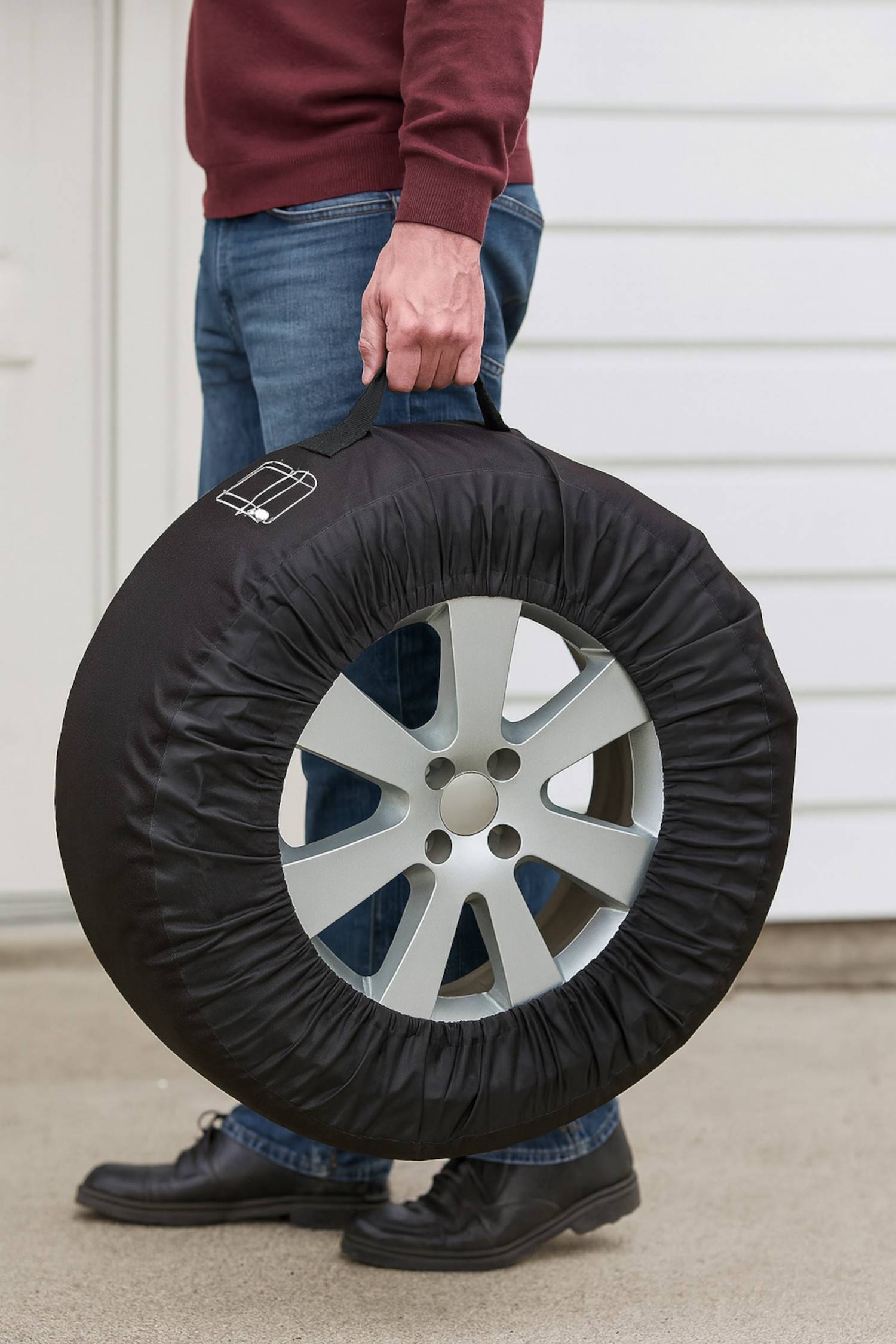 Eine Person, die ein Autoreifen mit einer schwarzen Schutzabdeckung hält, wobei ein silberner Alufelge teilweise sichtbar ist, vor einer weißen Wand.