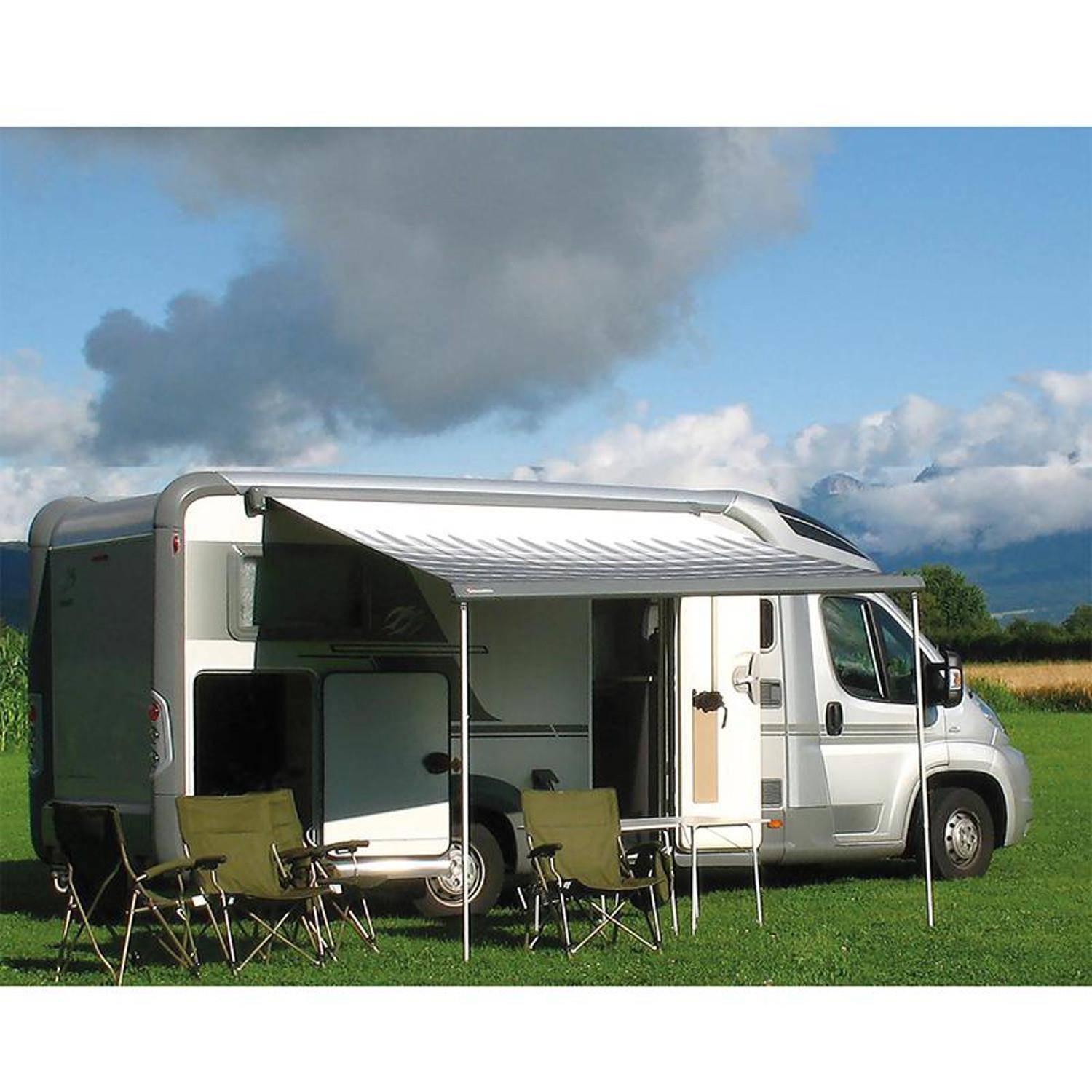 Ein Wohnmobil, das auf Gras geparkt ist, mit ausgefahrener Markise und vier Campingstühlen, vor einer malerischen Kulisse aus Wolken und Bergen.
