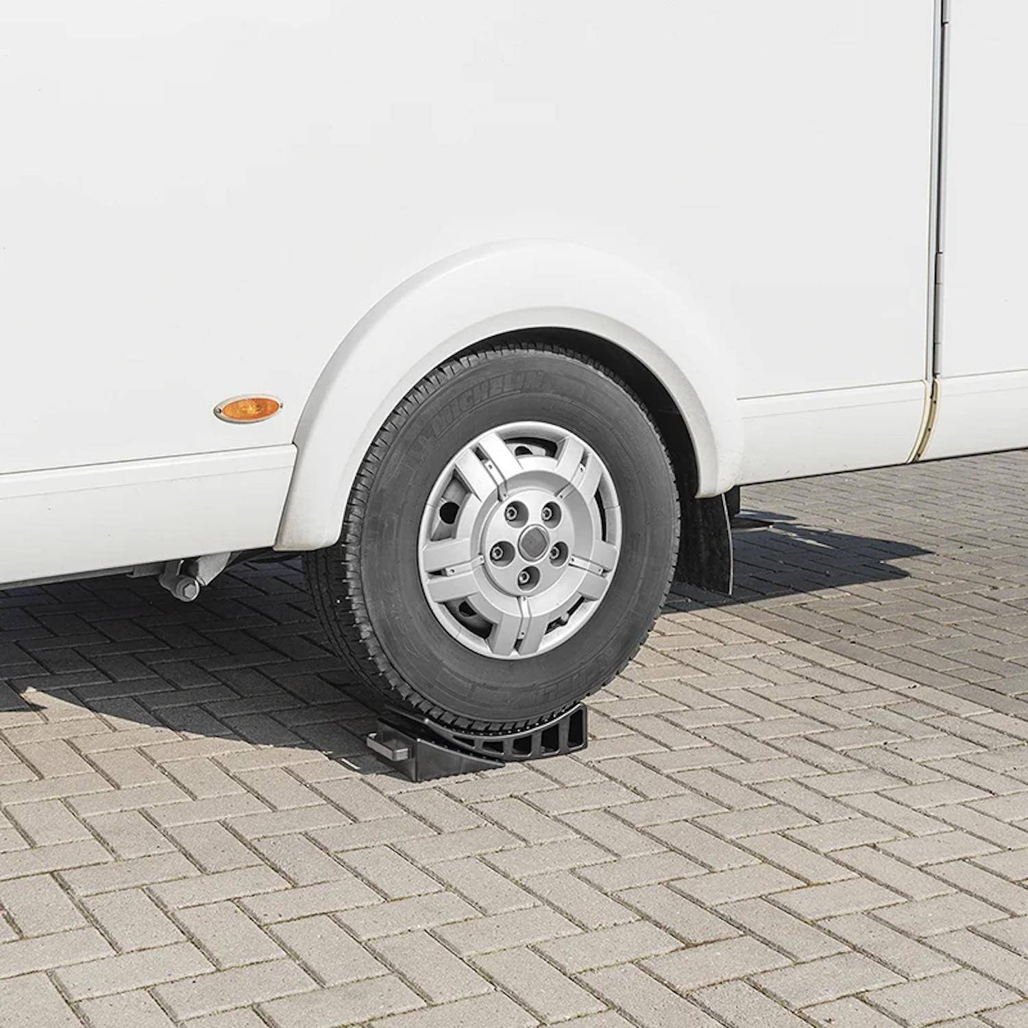 Ein weißes Fahrzeug hat sein Hinterrad auf einer schwarzen Kunststofframpe auf einem grauen Pflasterstein-Untergrund angehoben, höchstwahrscheinlich zur Wartung oder Ausrichtung.