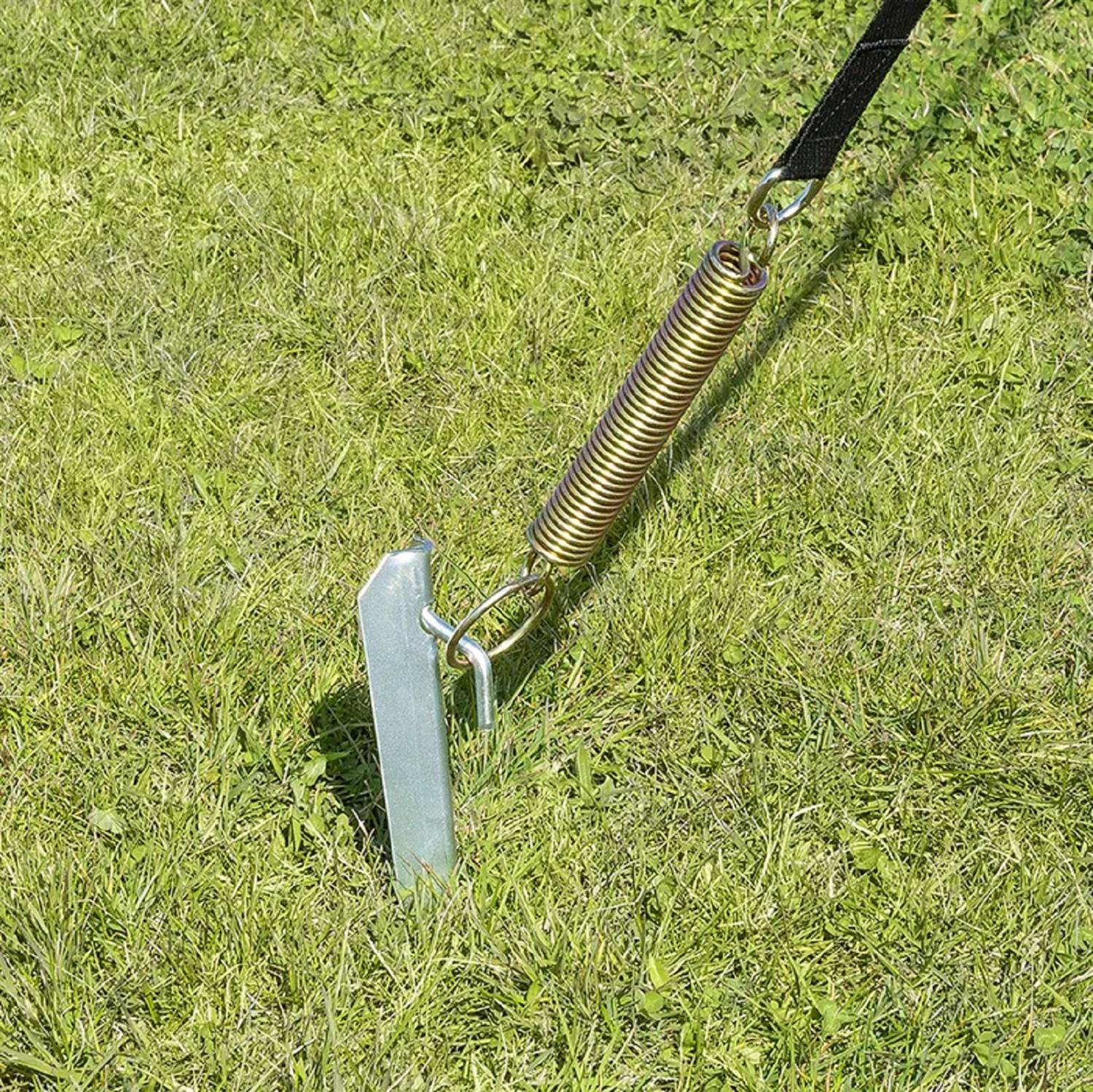 Ein Metallpfahl mit einer angebrachten Schraubenfeder wird in den grasigen Boden gerammt, wahrscheinlich um eine Konstruktion zu verankern oder eine Spannvorrichtung zu befestigen.