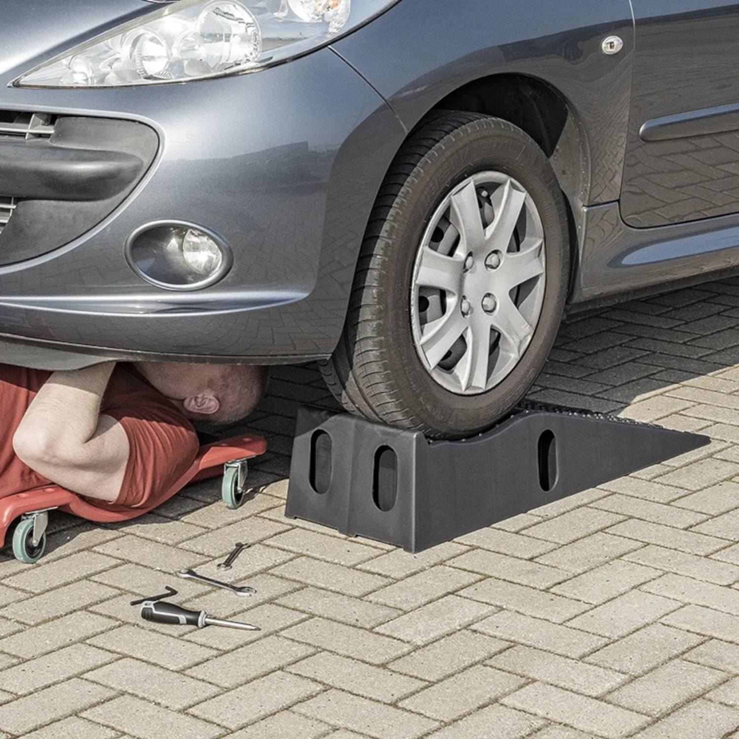 Auto auf Auffahrrampen in einem Parkplatz, während eine Person darunter Reparaturen durchführt. Ein Schraubenschlüssel und ein Schraubendreher liegen in der Nähe auf dem Boden.