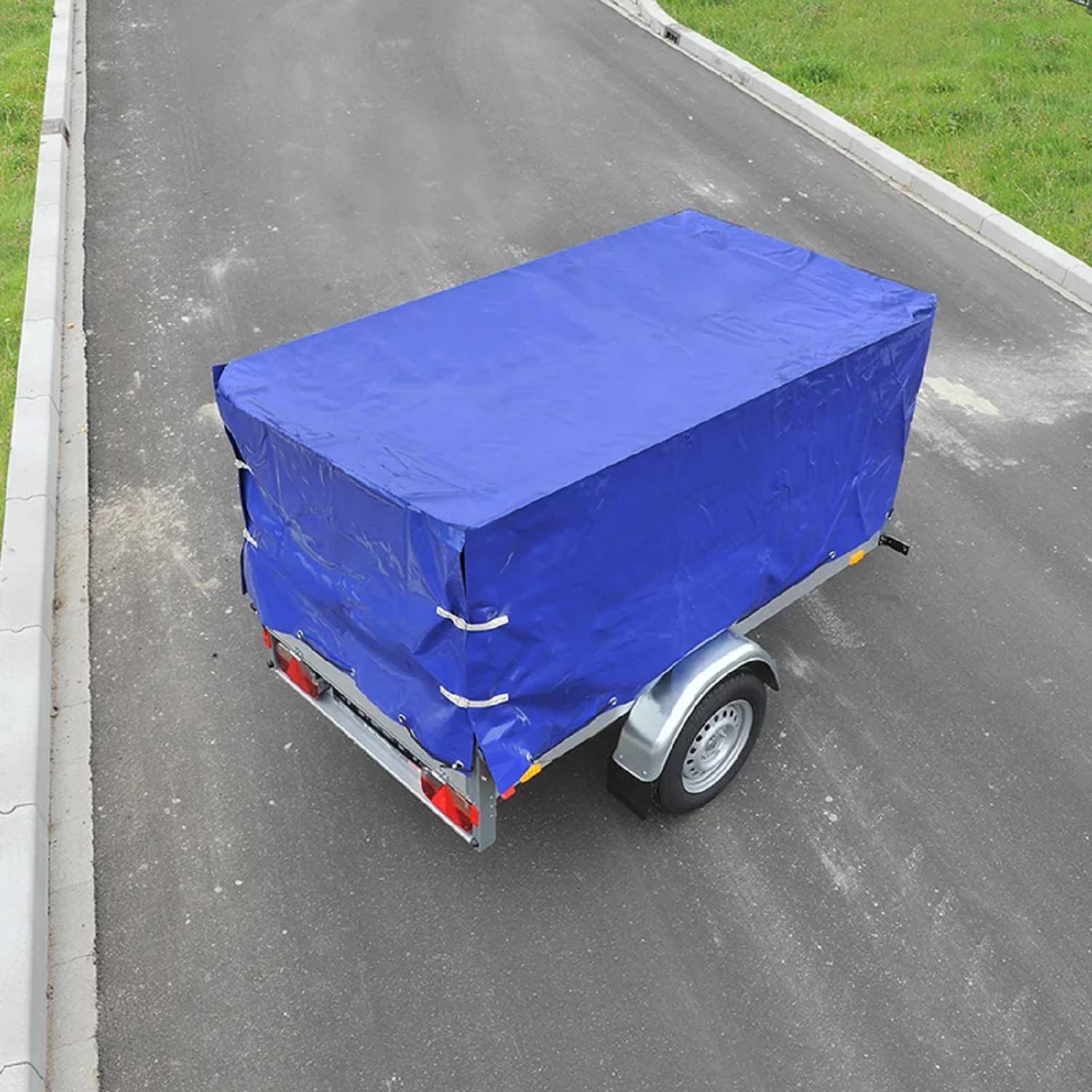 Ein kleiner Anhänger ist auf einer asphaltierten Straße geparkt, mit einer blauen Plane abgedeckt, neben einem Graßstreifen.