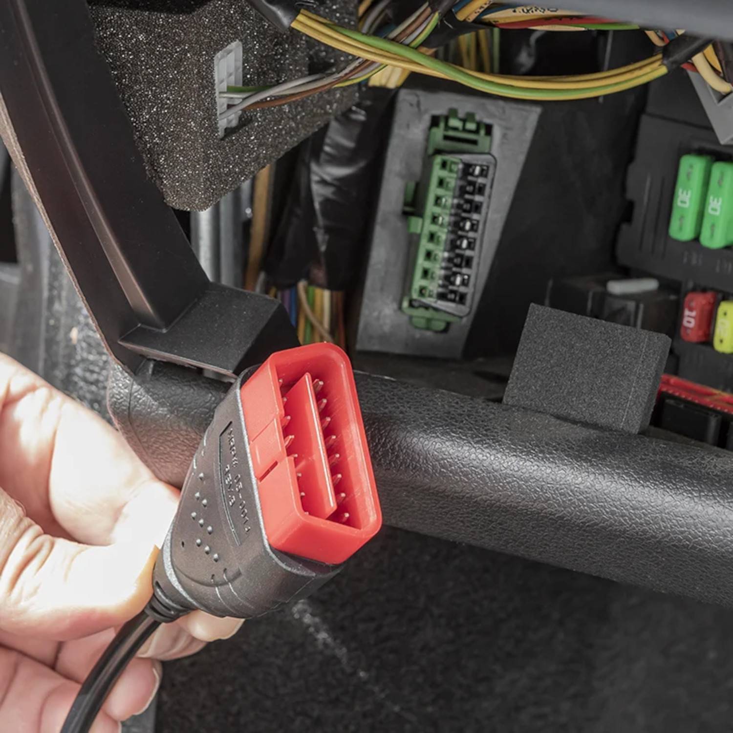 Eine Hand, die ein rotes Diagnosegerät in einen OBD2-Anschluss unter einem Armaturenbrett einsteckt, umgeben von Kabeln und anderen elektronischen Komponenten.