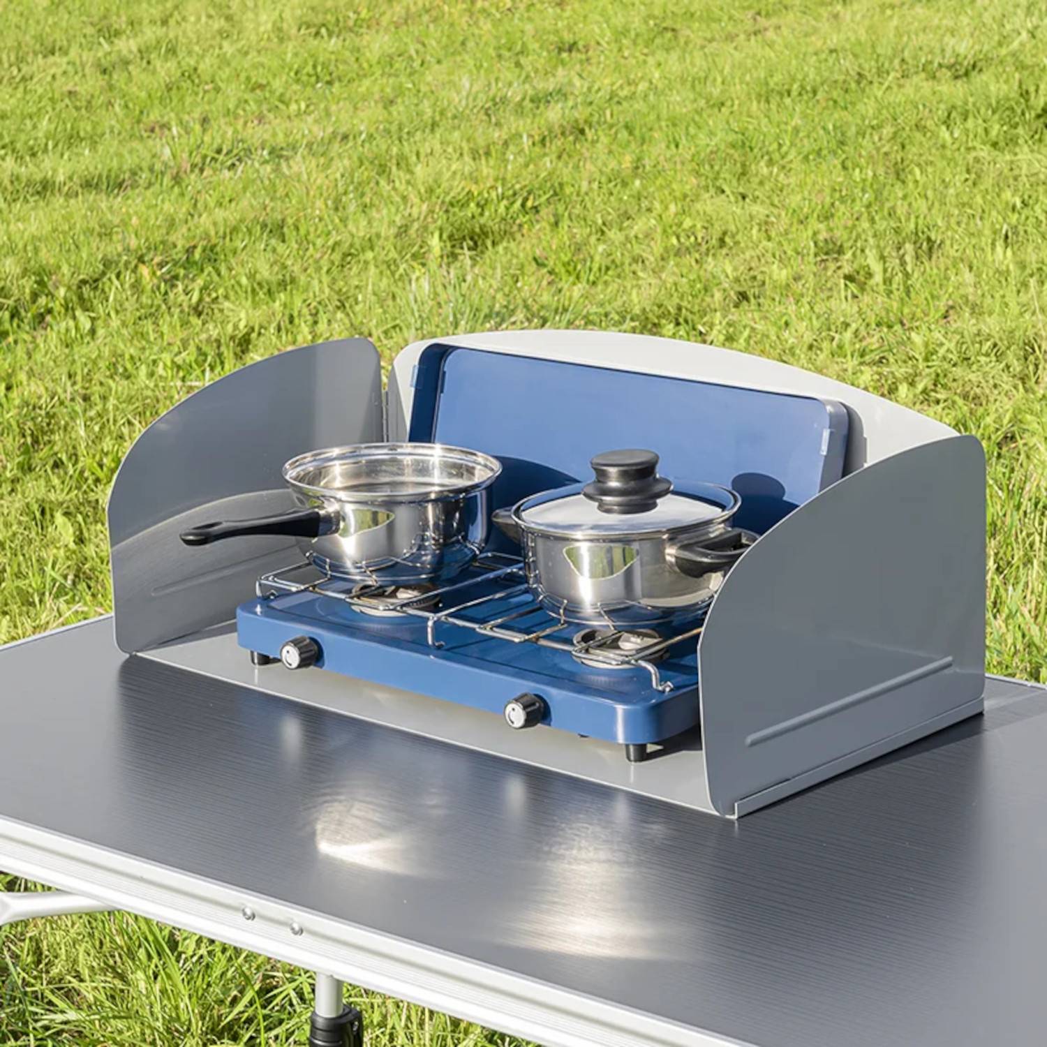 Ein tragbarer Campingkocher auf einem Tisch mit zwei Töpfen, die im Freien auf einem Rasenfeld kochen.