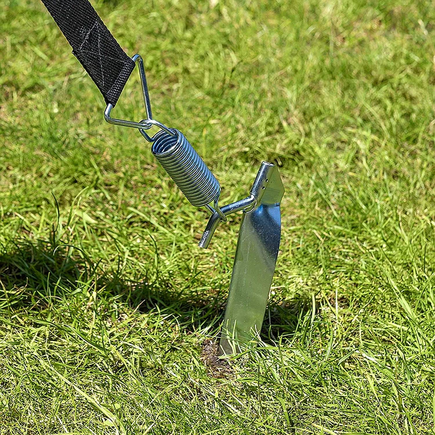 Ein Metallgartenpflock mit einer Feder und einem schwarzen Riemen verankert sich sicher im Rasen und ist darauf ausgelegt, Zelte oder Außenstrukturen zu stabilisieren.