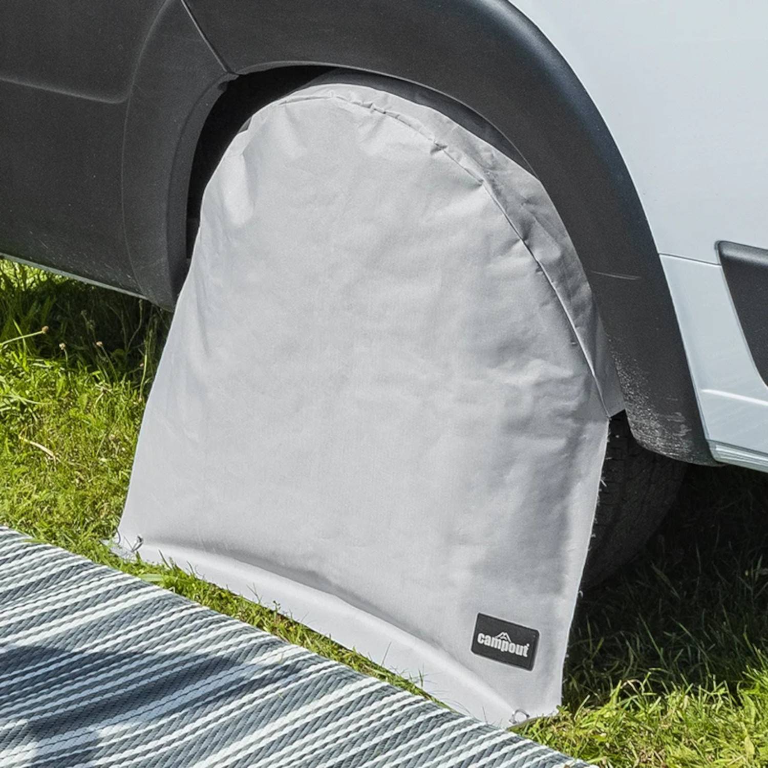 Eine Reifenabdeckung auf dem Rad eines geparkten Fahrzeugs, teilweise auf Gras, mit einem kleinen Abschnitt einer gestreiften Matte. Die Abdeckung enthält ein 'cam, out' Logo.