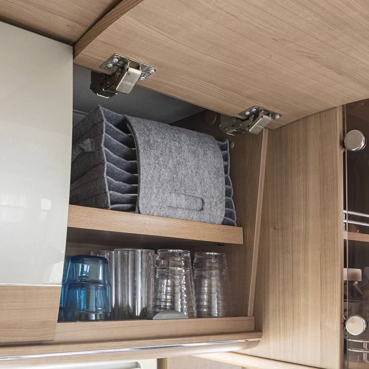 Oberer Küchenschrank mit Holzregalen, der ordentlich gestapelte graue Filzuntersetzer und klare Gläser enthält und damit eine organisierte Aufbewahrung präsentiert.