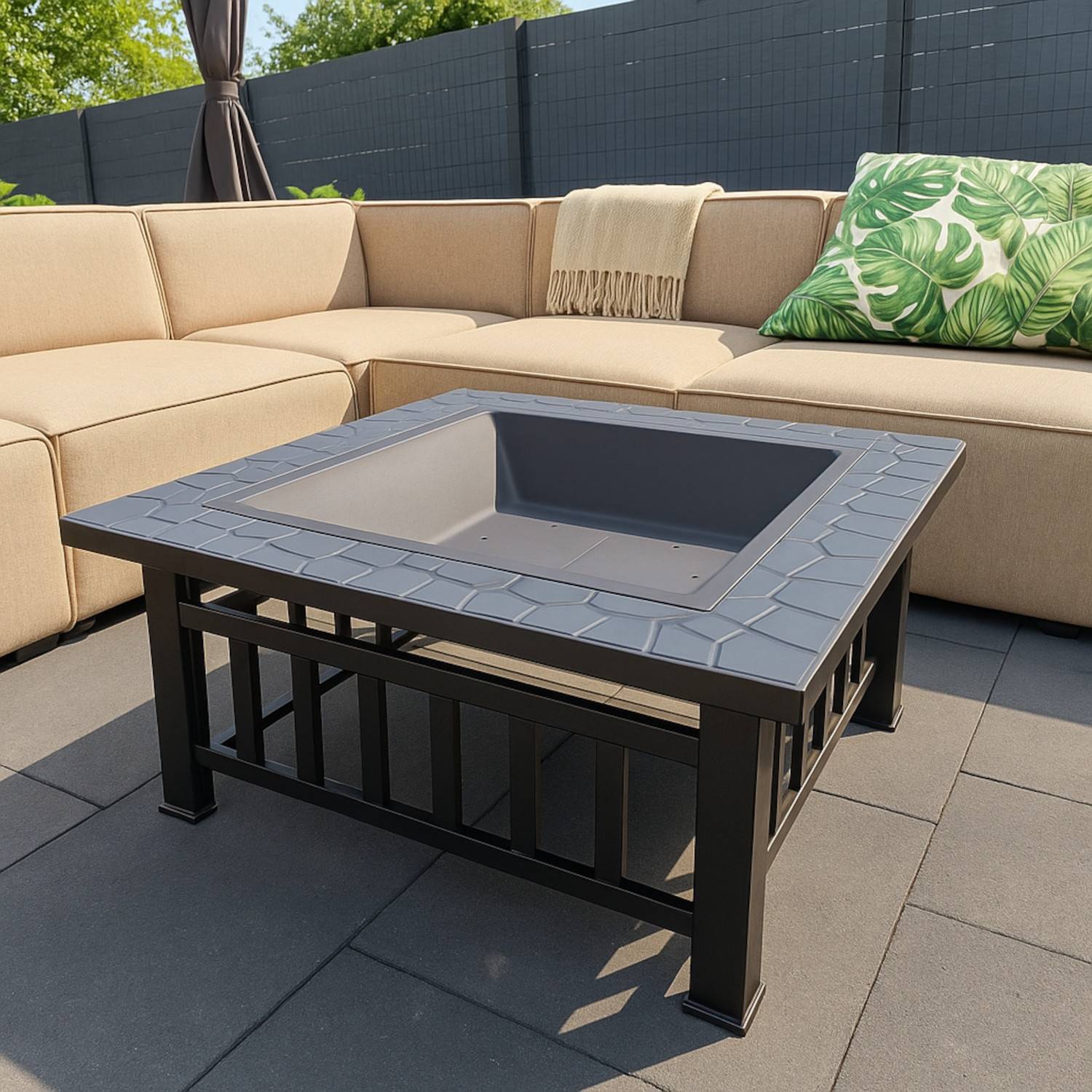 Eine beige L-förmige Outdoor-Couch mit grünen Blatt-gemusterten Kissen ist um einen schwarzen Metallfeuertisch auf einer gefliesten Terrasse angeordnet.