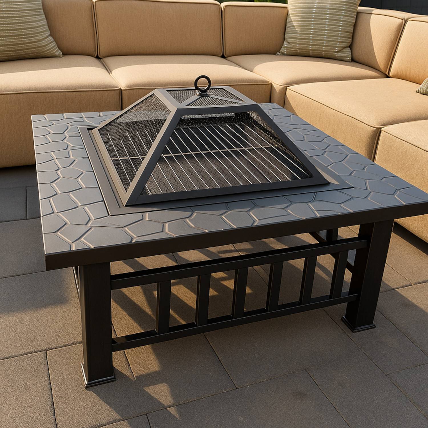 Ein quadratischer Outdoor-Feuertisch mit einem Metallgitterdeckel steht auf einer Terrasse und wird von einem beigen Ecksofa mit grün gestreiften Kissen umgeben.