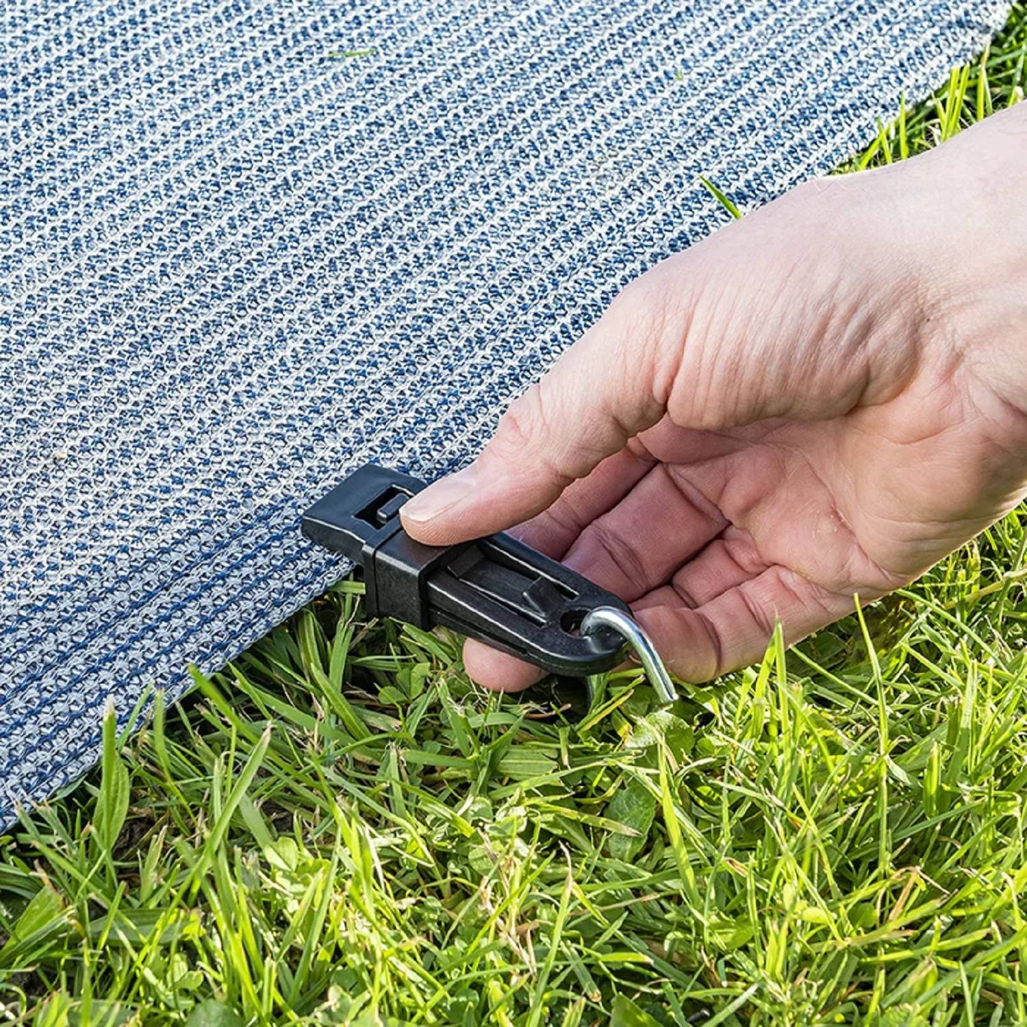 Eine Hand befestigt einen blau-weiß strukturierten Teppich auf Gras mit einer schwarzen Klammer und einem Pflöck, um Bewegung zu verhindern.