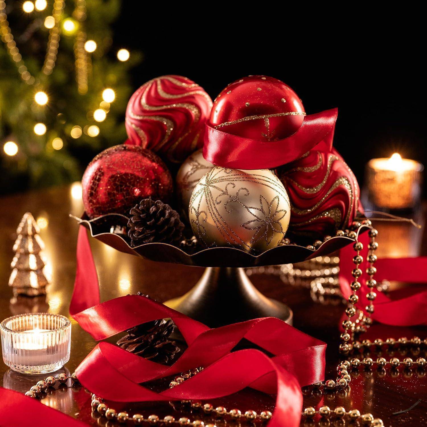 Eine festliche Anordnung zeigt rote und goldene Christbaumkugeln in einer Schale, dekoriert mit rotem Band und Perlengirlande, vor einem Hintergrund von brennenden Kerzen und einem Weihnachtsbaum.