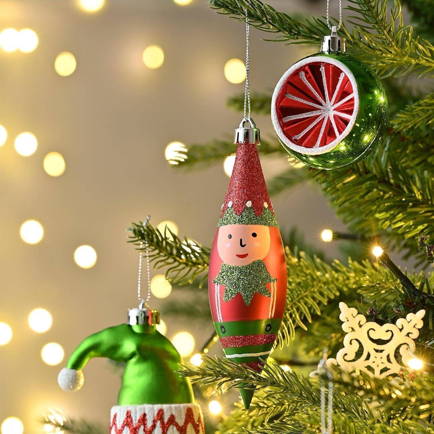 Weihnachtsbaum mit dekorativen Schmuckstücken, einschließlich einer grünen Weihnachtskugel, einer rot-grünen Elfenaufhängung und Lichtern, die eine festliche Atmosphäre erzeugen.