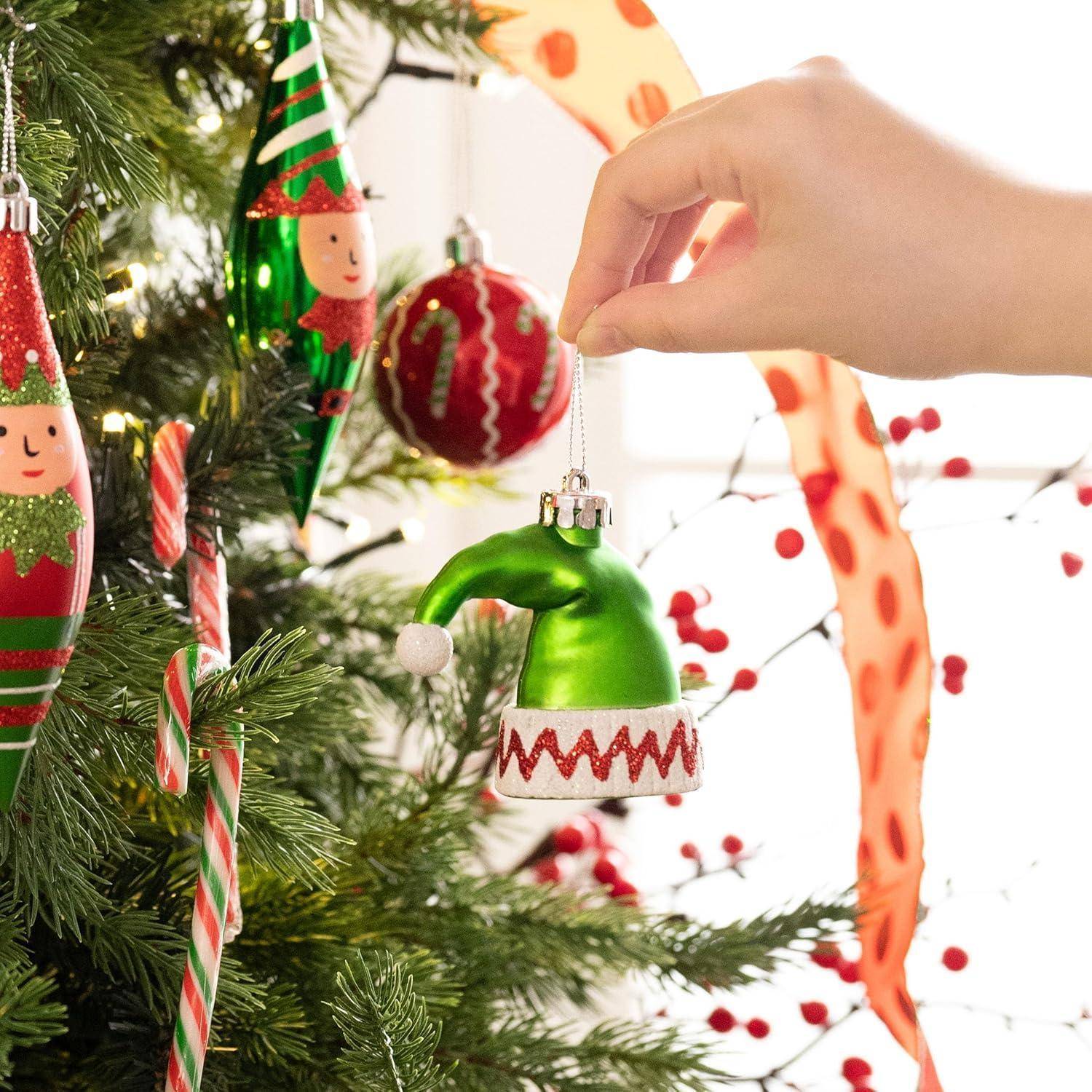 Eine Hand platziert einen grünen Elfenhut-Anhänger auf einem Weihnachtsbaum, der mit Zuckerstangen-Dekorationen und verschiedenen festlichen Ornamenten geschmückt ist.