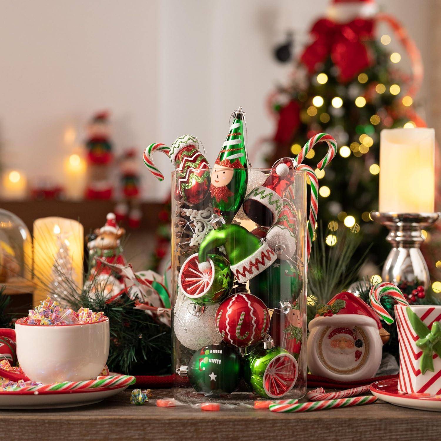 Festlich gedeckter Feiertagstisch mit einer Glasvase voller bunter Christbaumkugeln und Zuckerstangen, umgeben von Kerzen und einem unscharf im Hintergrund stehenden Weihnachtsbaum.