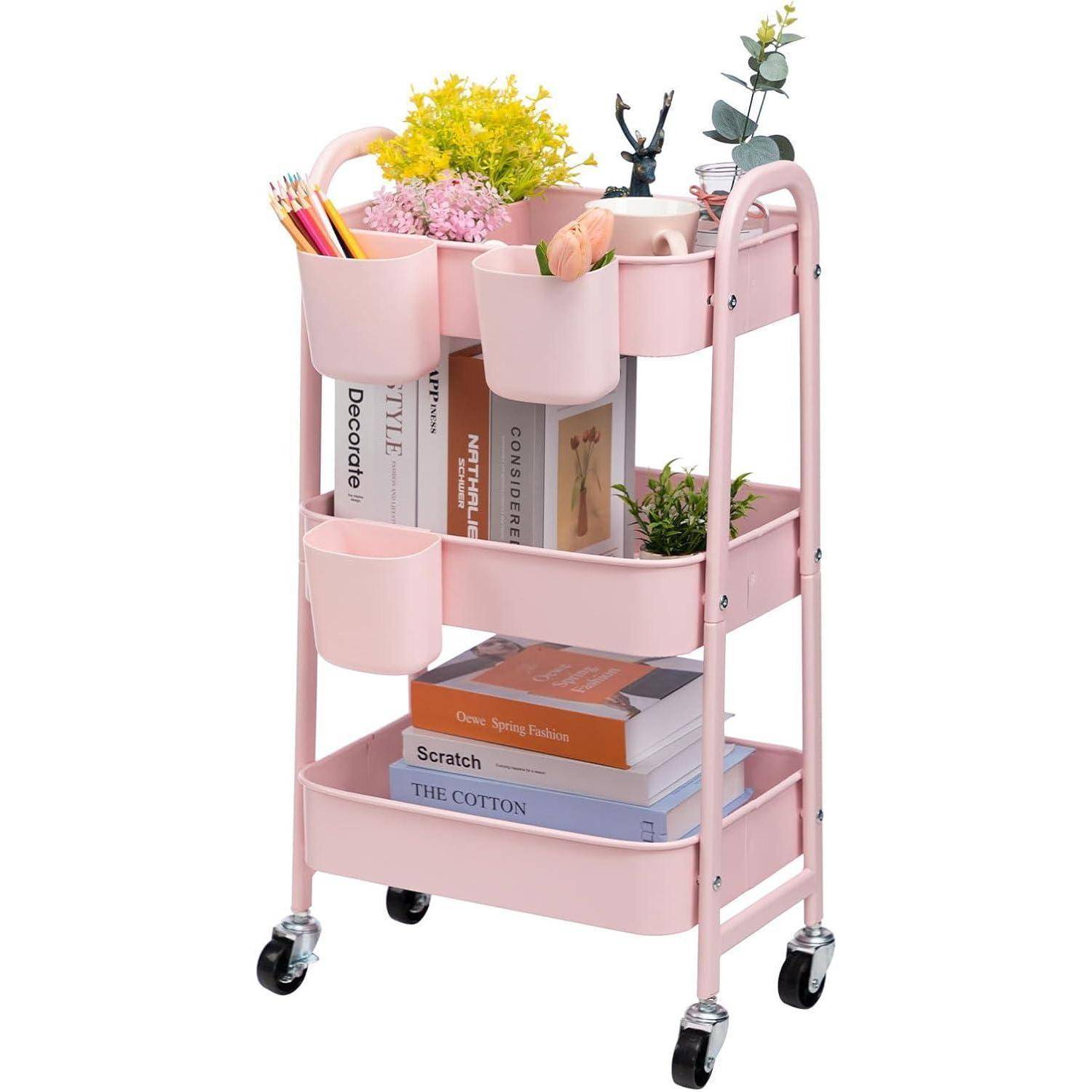 Ein rosa rollbarer Wagen mit drei Regalen, der Bücher, Pflanzen und Schreibwaren wie Scheren und Stifte effizient organisiert trägt.