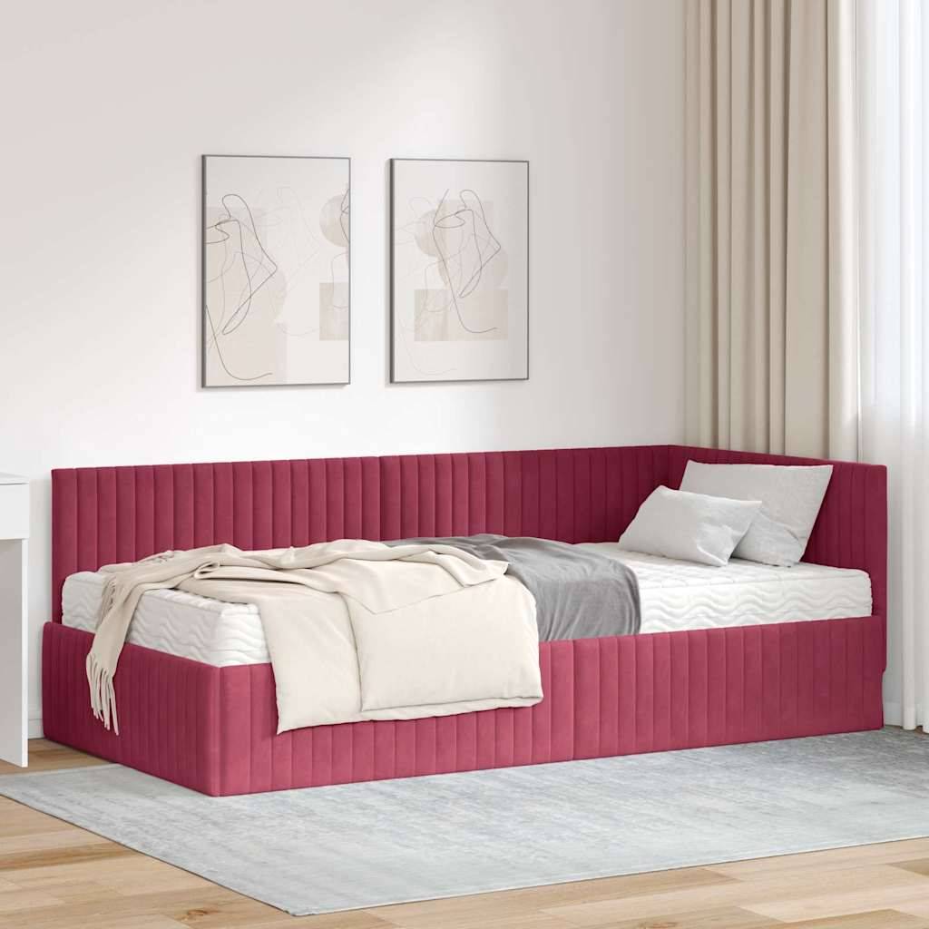 Ein modernes Schlafzimmer zeigt ein L-förmiges Bett in tiefdunkelrot mit weißer Bettwäsche und grauen Kissen, das vor einer helltonigen Wand mit abstrakter Kunst platziert ist.