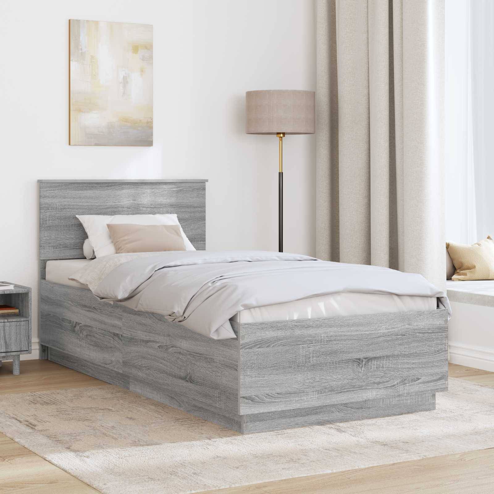 Ein modernes Schlafzimmer zeigt ein Bett aus grauem Holz mit weißer und grauer Bettwäsche, einen passenden Nachttisch und eine hohe beige Lampe neben cremefarbenen Gardinen.