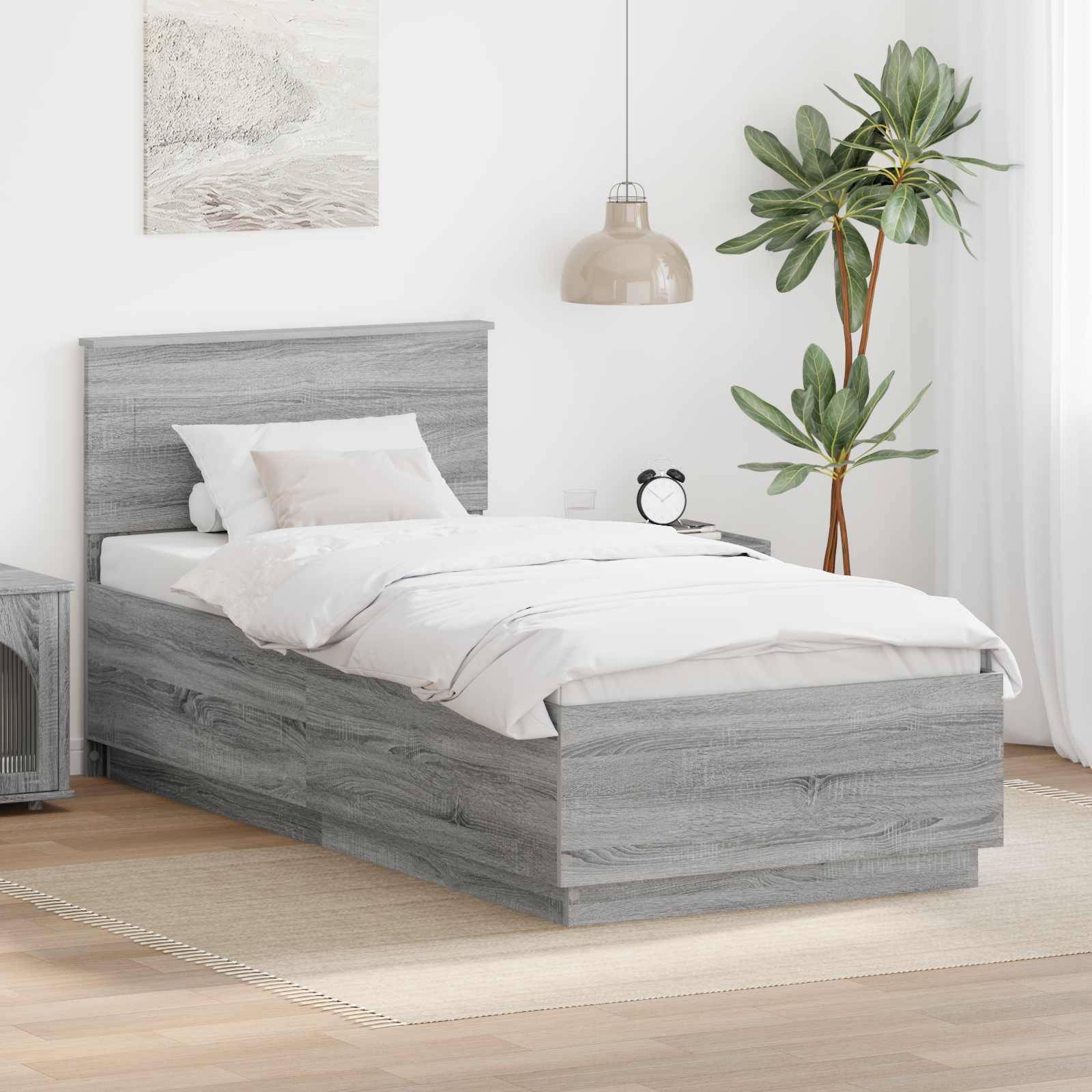Ein ordentlich gemachtes Bett mit grauem Holzrahmen und weißer Bettwäsche in einem hellen Schlafzimmer, mit einem kleinen Nachttisch, einer hängenden Lampe und einer Topfpflanze.