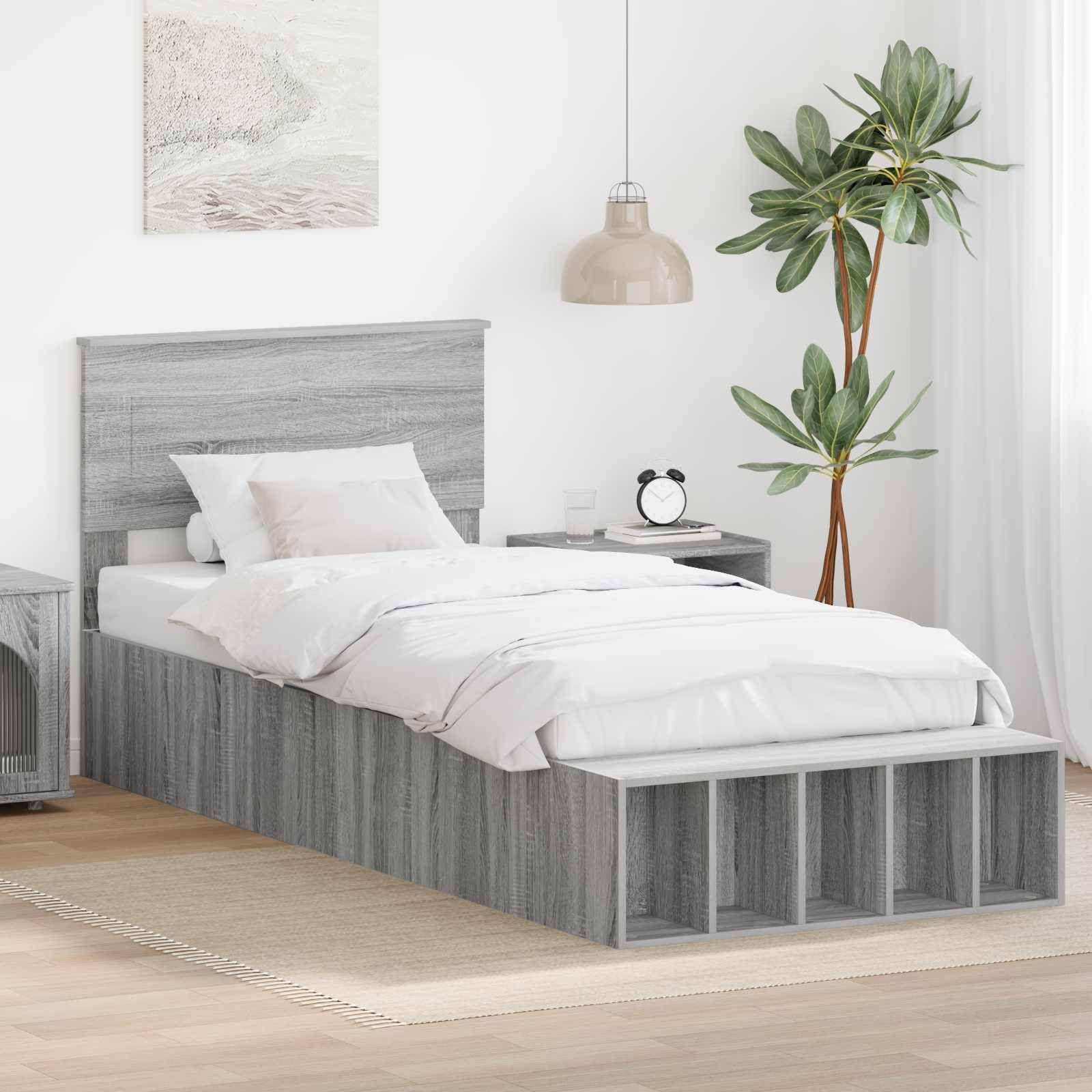 Ein ordentlich gemachtes Einzelbett mit grauem Holzrahmen und weißer Bettwäsche. Eine Topfpflanze, eine hängende Lampe und ein kleiner Nachttisch befinden sich in der Nähe.