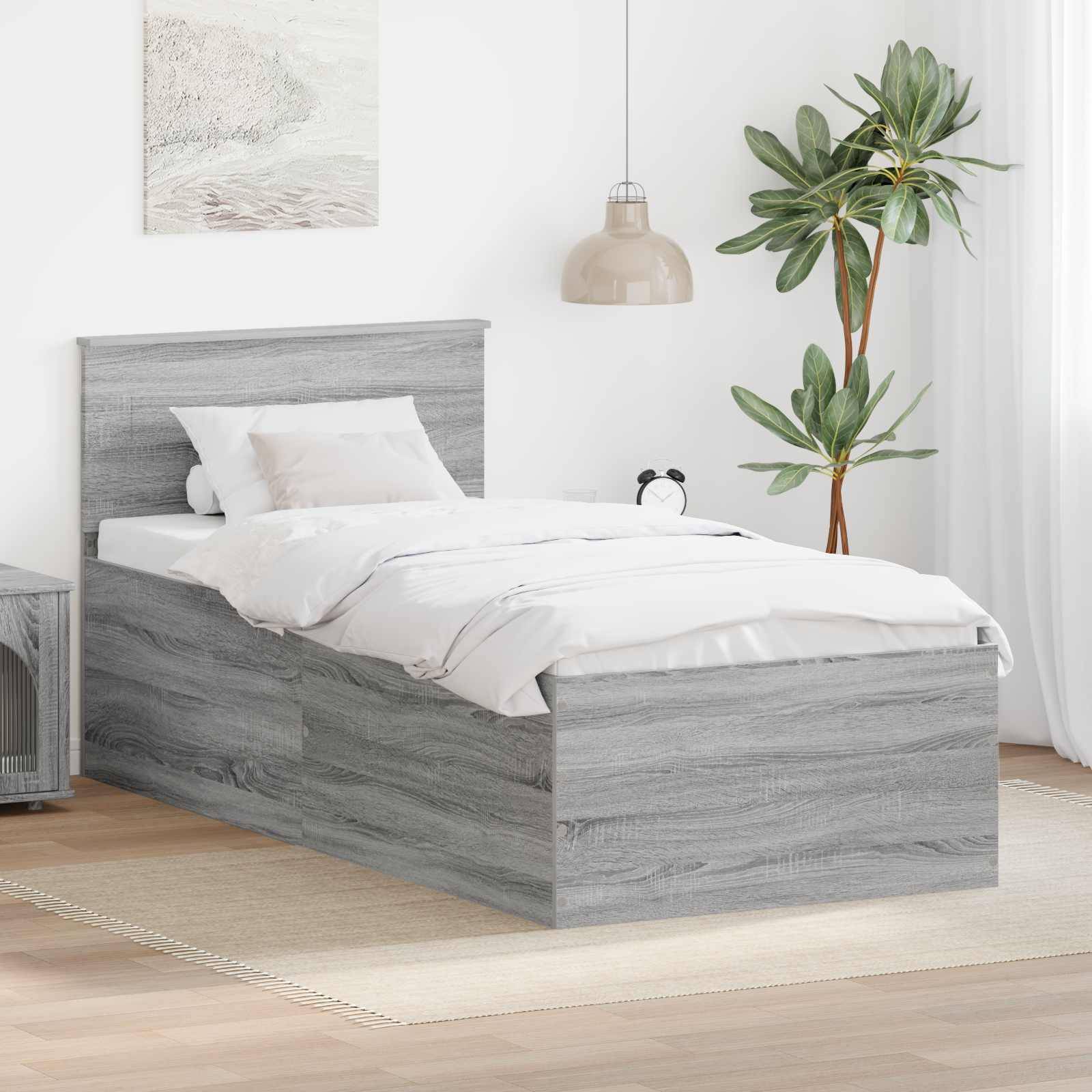 Ein grauer Holzbett mit weißer Bettwäsche steht in einem hellen Raum, der eine grüne Pflanze, Wandkunst und eine moderne Pendelleuchte aufweist.