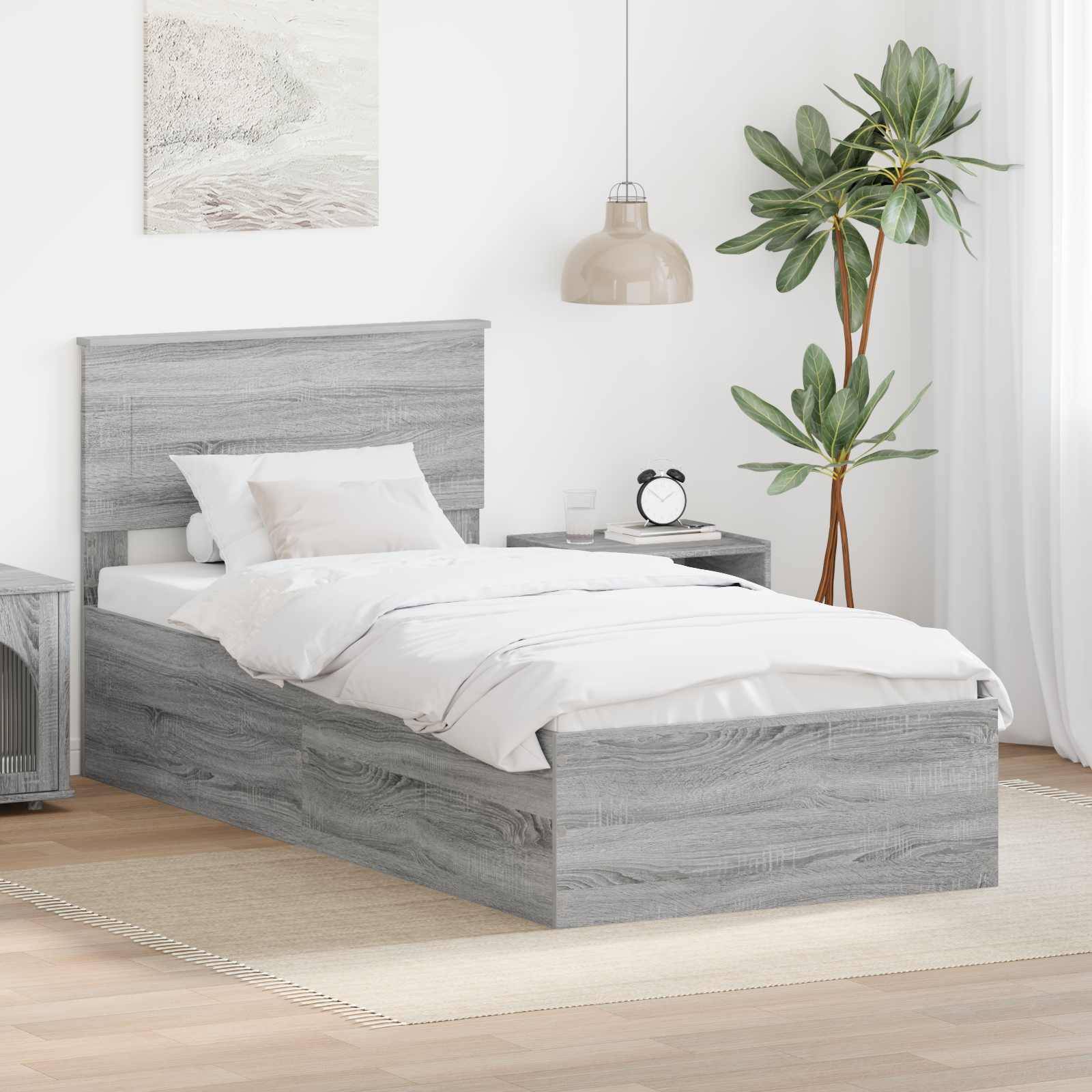 Modernes Schlafzimmer mit einem Bett aus grauem Holz, weißer Bettwäsche, einem Nachttisch mit einer Lampe und einem Wecker sowie einer hohen Topfpflanze in der Ecke.