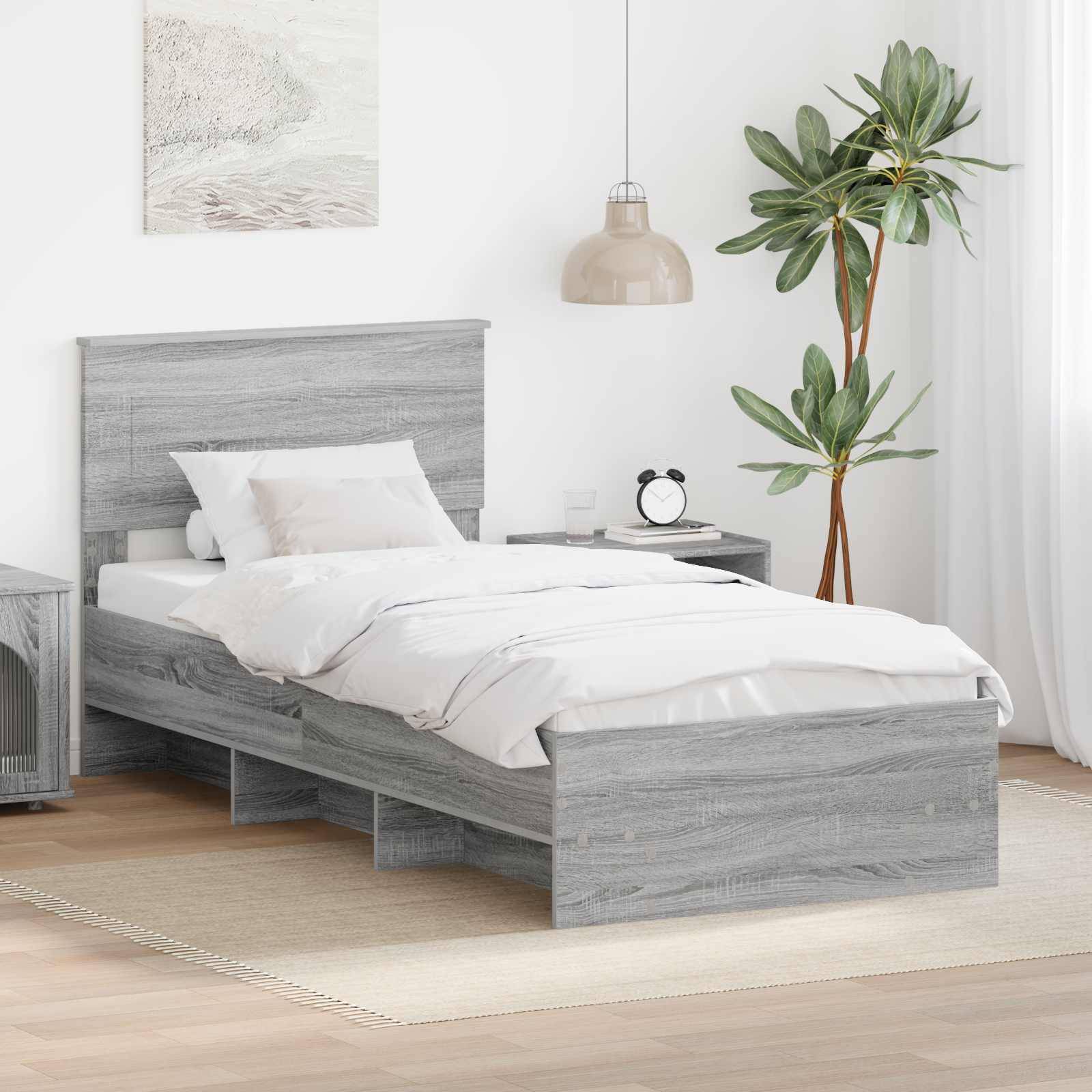 Ein modernes Schlafzimmer mit einem grauen Holzbett, weißer Bettwäsche, Kissen und einem passenden Nachttisch. Eine Lampe und eine Pflanze dienen als Dekoration.