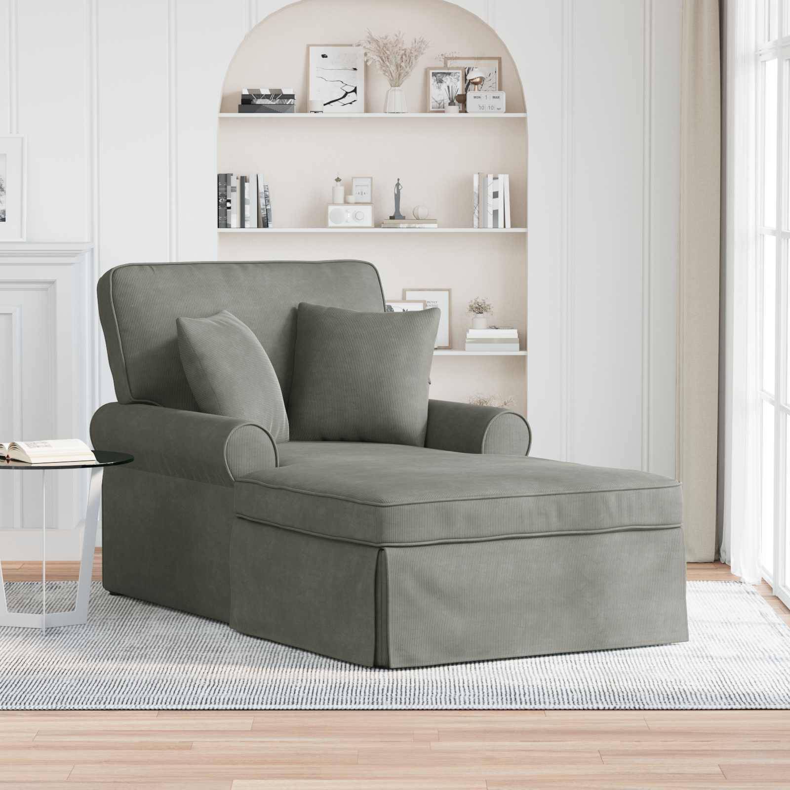 Ein grauer Sessel mit zwei Kissen steht auf einem hellen Teppich in einem Wohnzimmer. Eine weiße Wand zeigt ein eingebautes Regal mit Büchern und Dekorationsstücken.