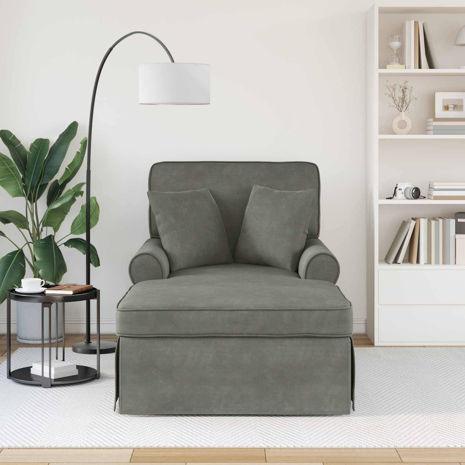 Ein grauer Sessel mit zwei Kissen steht im Wohnzimmer neben einer Stehlampe, einem kleinen runden Tisch mit Pflanzen und einem weißen Bücherregal.