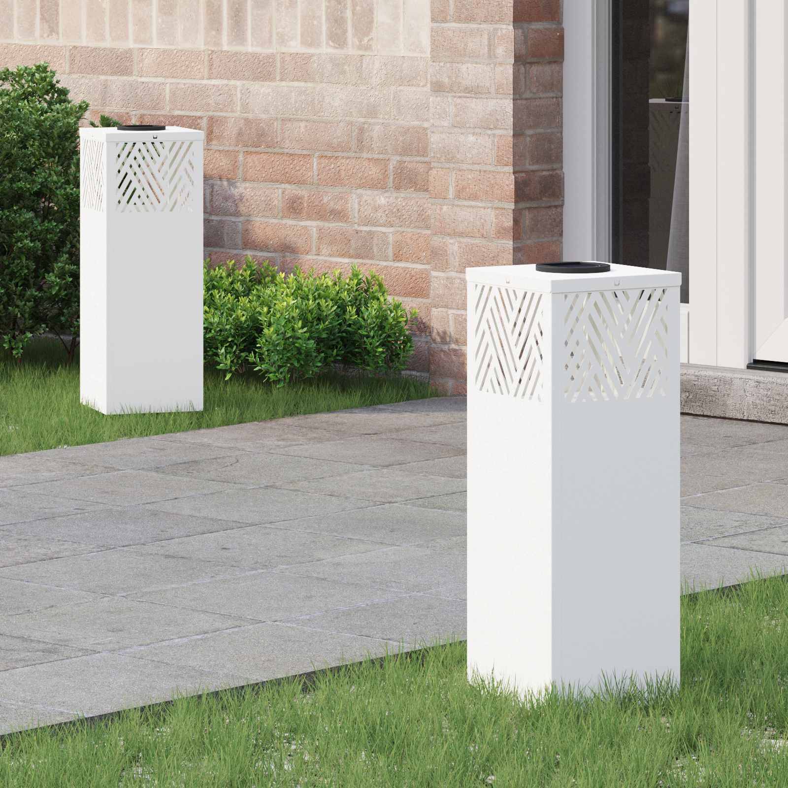 Zwei weiße Außensäulen mit dekorativen Ausschnitten stehen auf einem gefliesten Weg, umgeben von Gras und kleinen Sträuchern in der Nähe einer Ziegelwand.