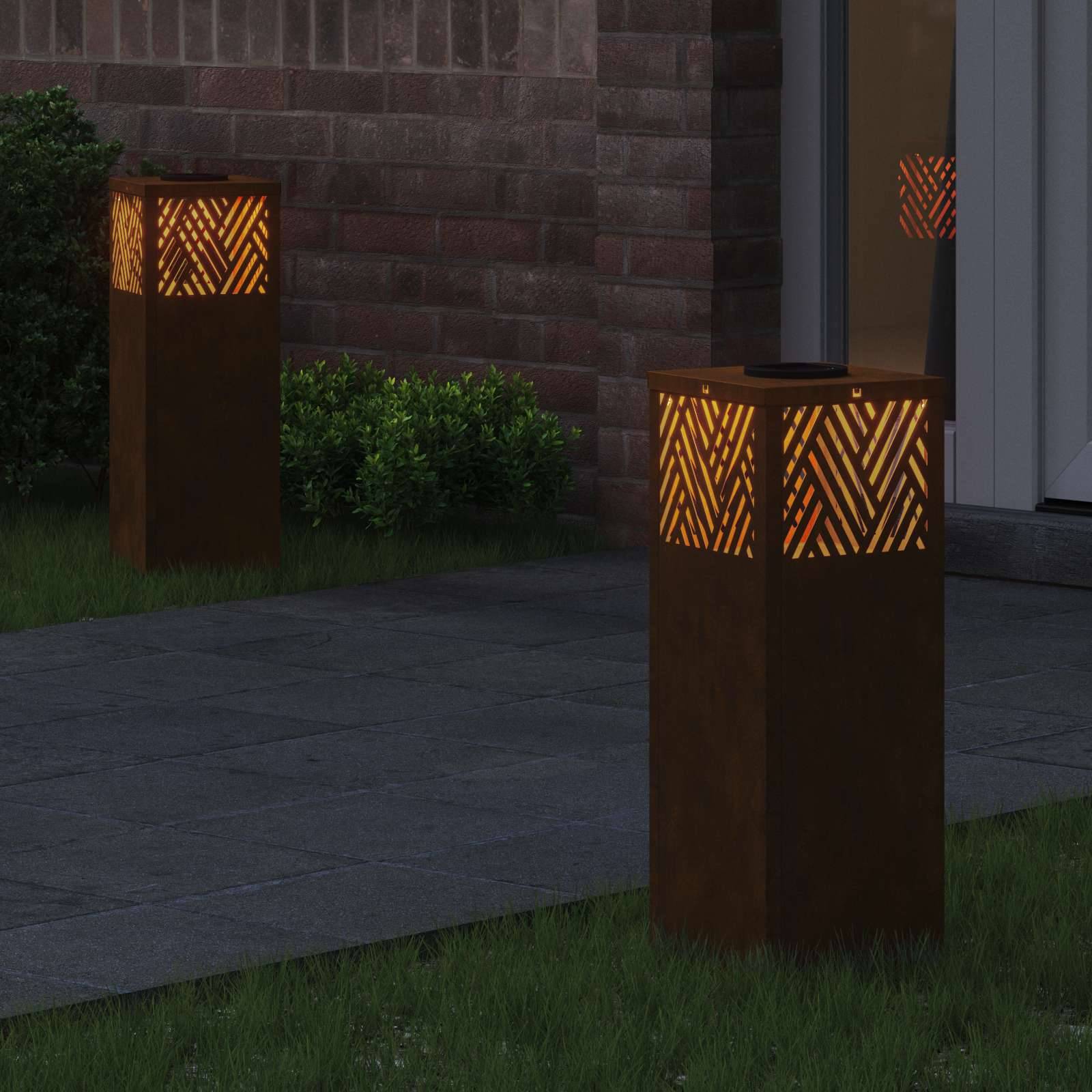 Zwei Außenlampen mit geometrischen Mustern verbreiten warmes Licht und beleuchten einen Gartenweg neben einer Ziegelmauer und grünen Sträuchern.