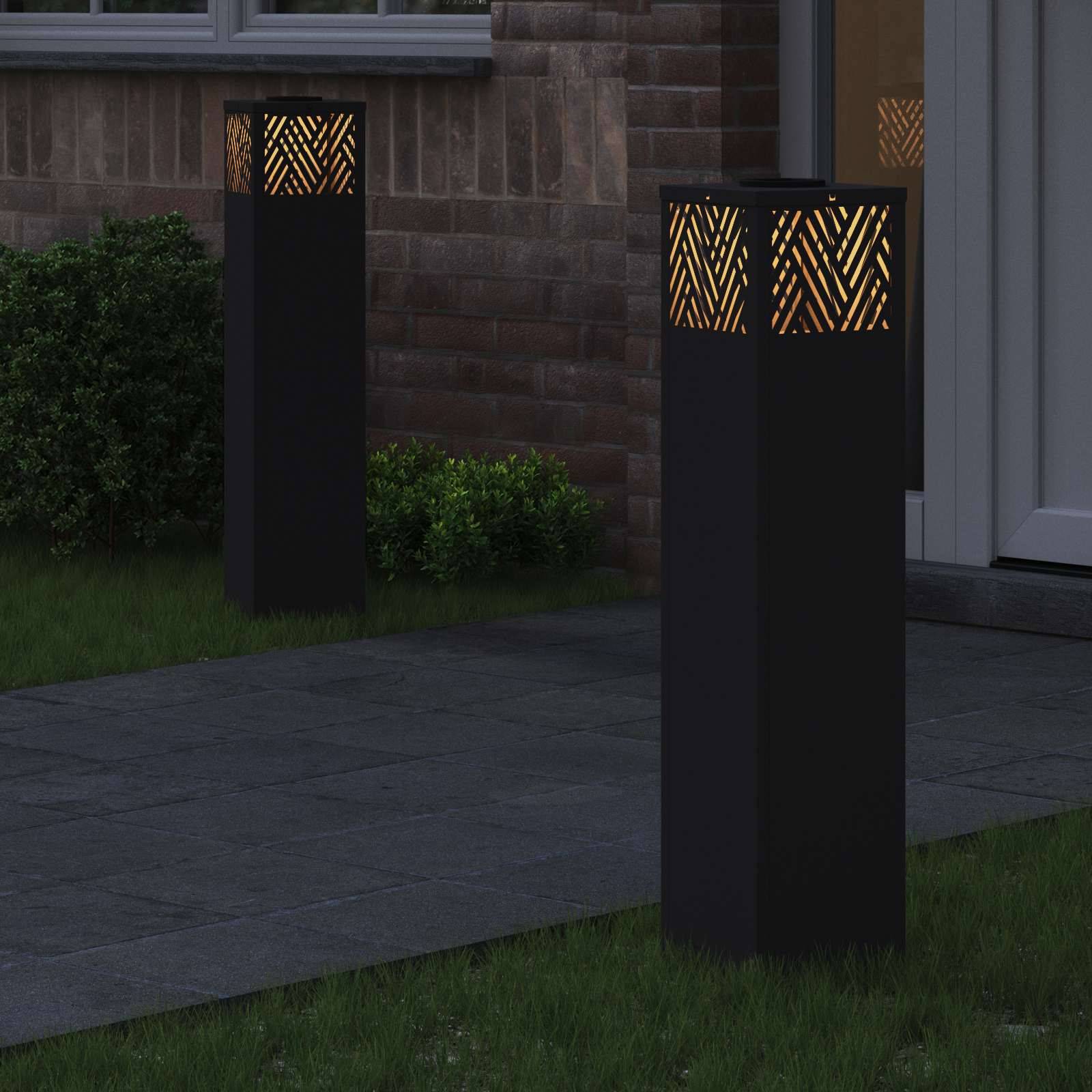 Ein schwach beleuchteter Weg wird von zwei modernen, schwarzen, quadratischen Lampen mit dekorativen Ausschnitten gesäumt, die zu einem Backsteingebäude führen.