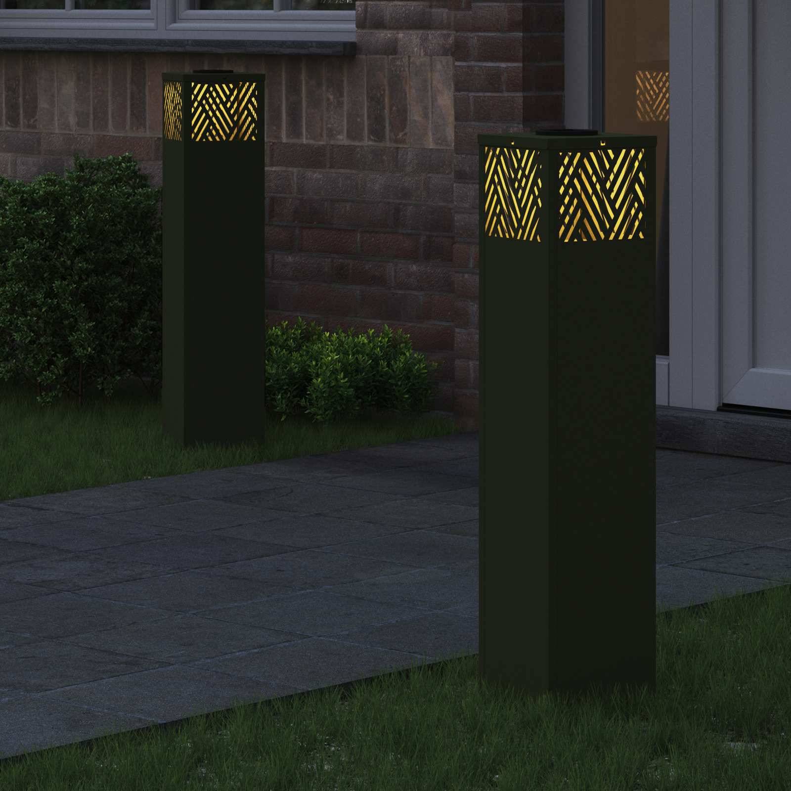 Zwei moderne Außenlampenmasten mit geometrischen Ausschnitten strahlen warmes Licht in einem Garten mit Pflastersteinen und einer Ziegelgebäude-Kulisse.