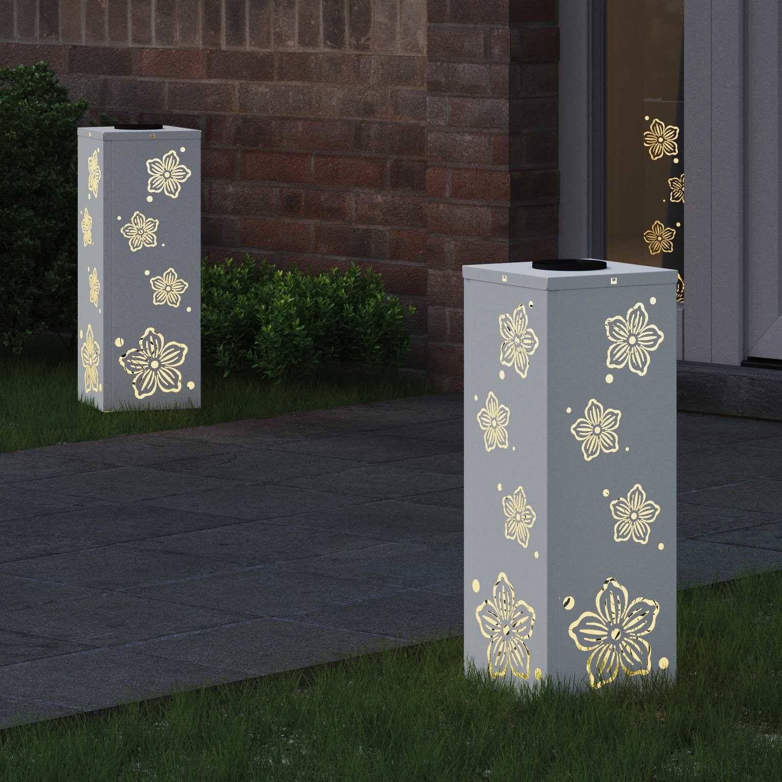 Zwei dekorative Gartenlampen mit floralen Ausschnitten verbreiten ein warmes Leuchten auf einem Steinweg neben einer Ziegelmauer und einer Glastür.