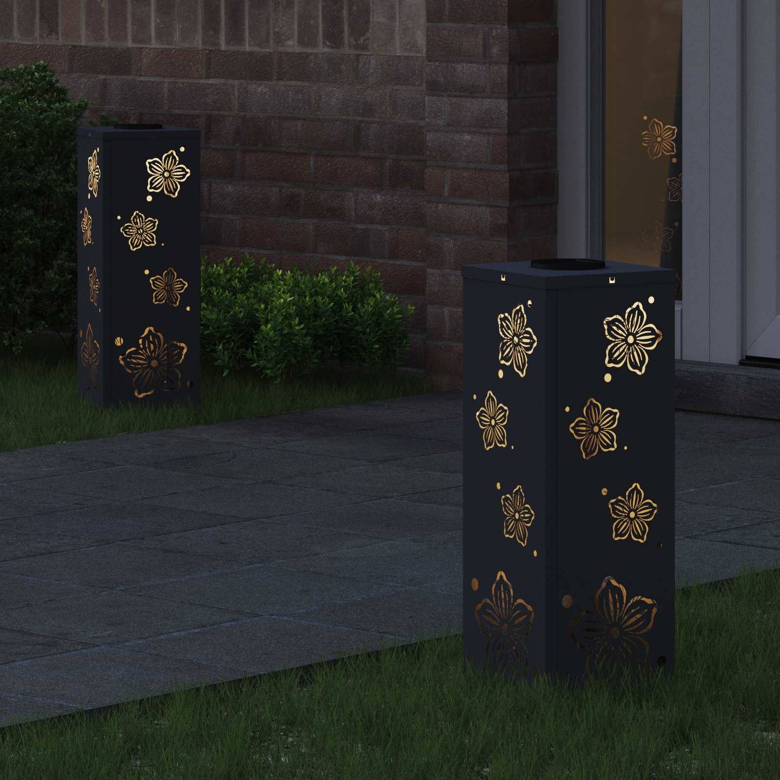 Ein Gartenweg wird von rechteckigen Laternen mit Blumenausschnitten beleuchtet, die gemusterte Schatten auf den Boden werfen, in der Nähe einer Backsteinmauer und Pflanzen.